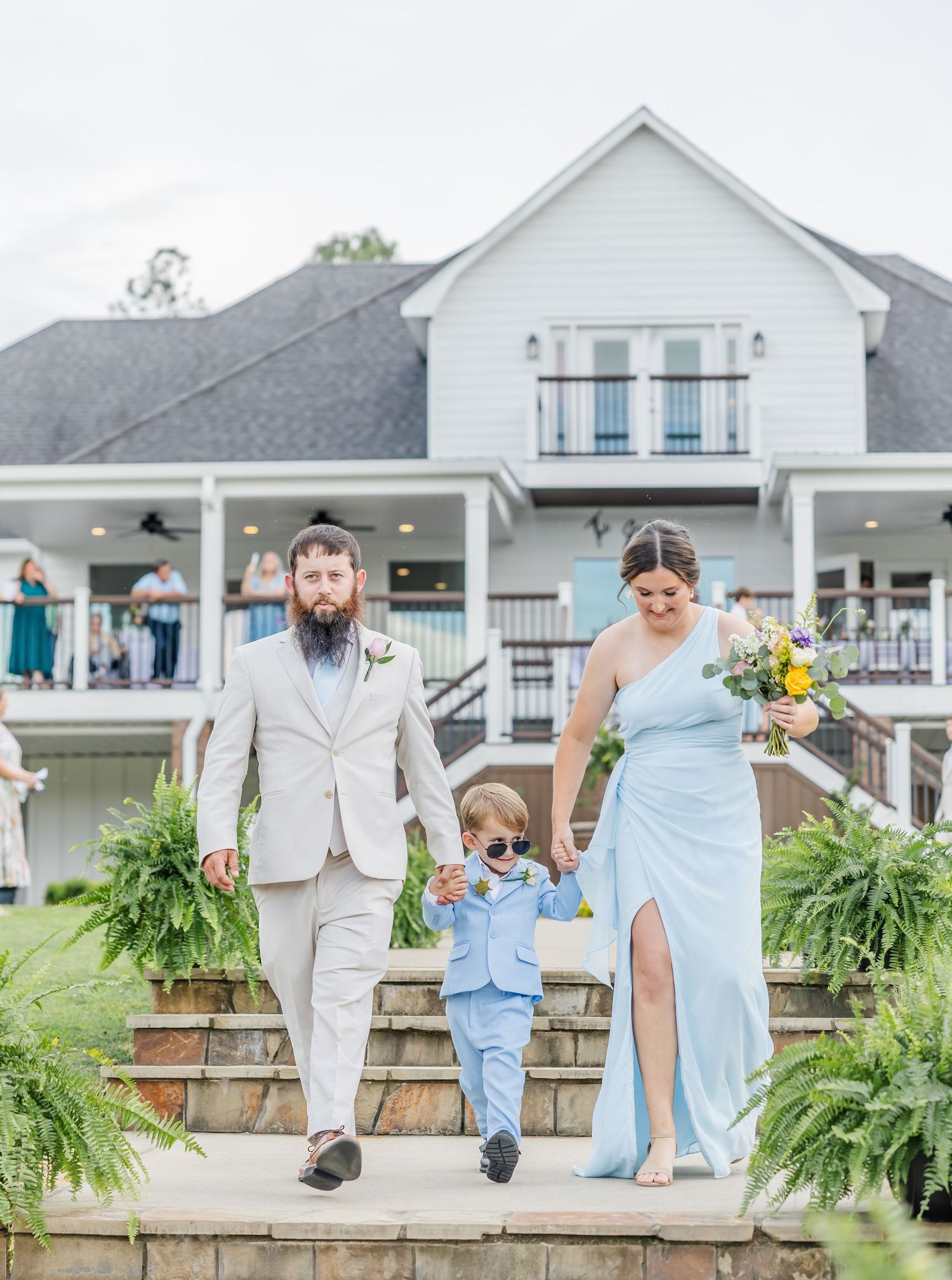 Bearded groomsman and woman in blue gown walking with young boy in sunglasses down venue steps.