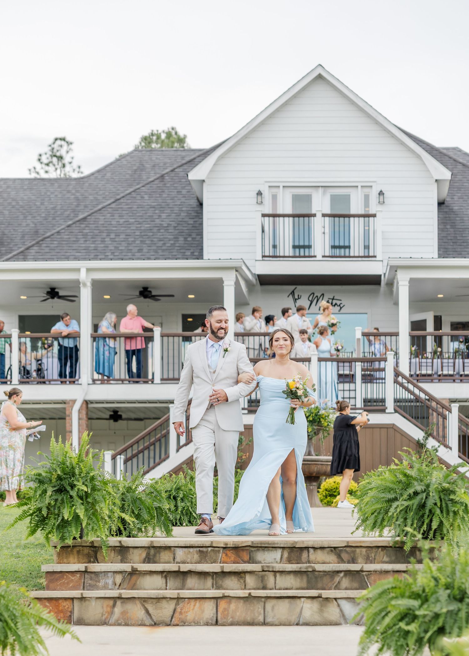 Smiling groom in tan suit escorts bridesmaid in blue gown down stone steps at outdoor wedding.