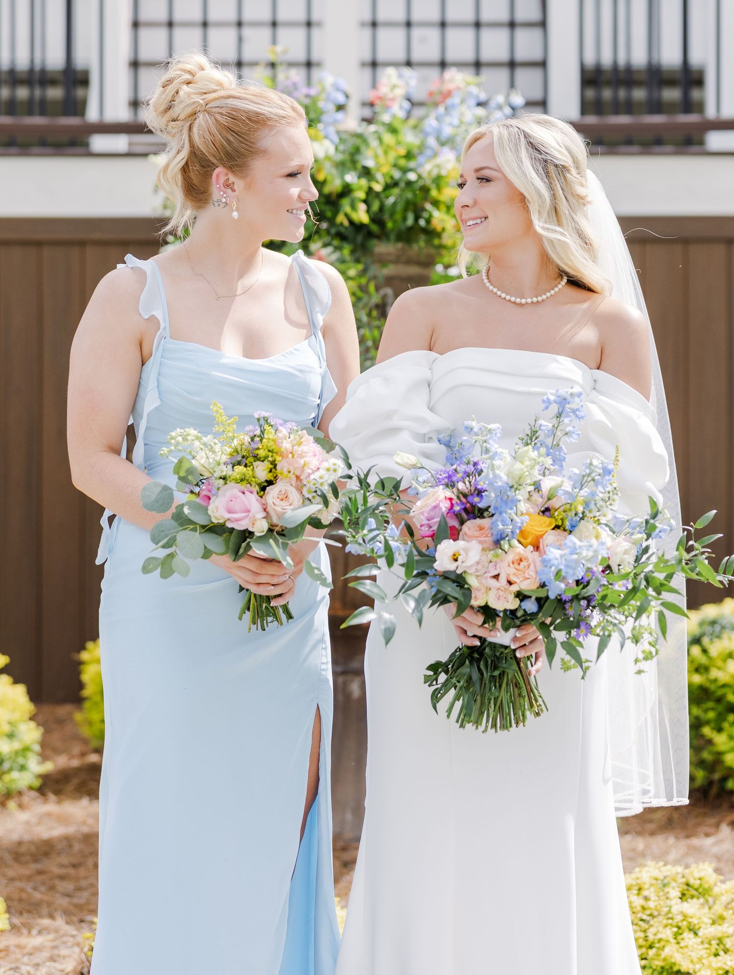 Bride in white off-shoulder gown and bridesmaid in light blue dress smile at each other holding bouquets.