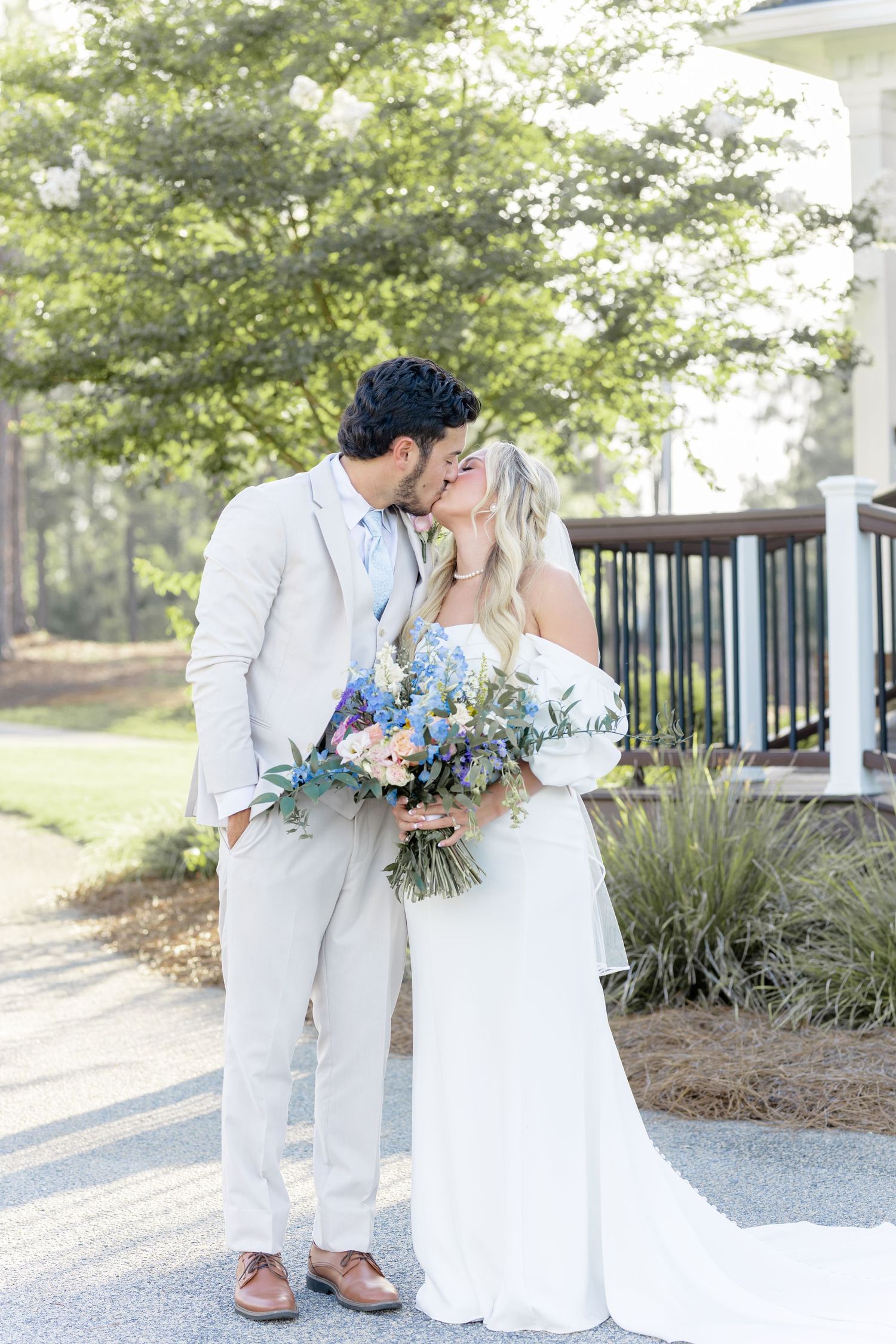 Bride and groom share a kiss while holding a blue and pink bouquet outdoors at their wedding venue.