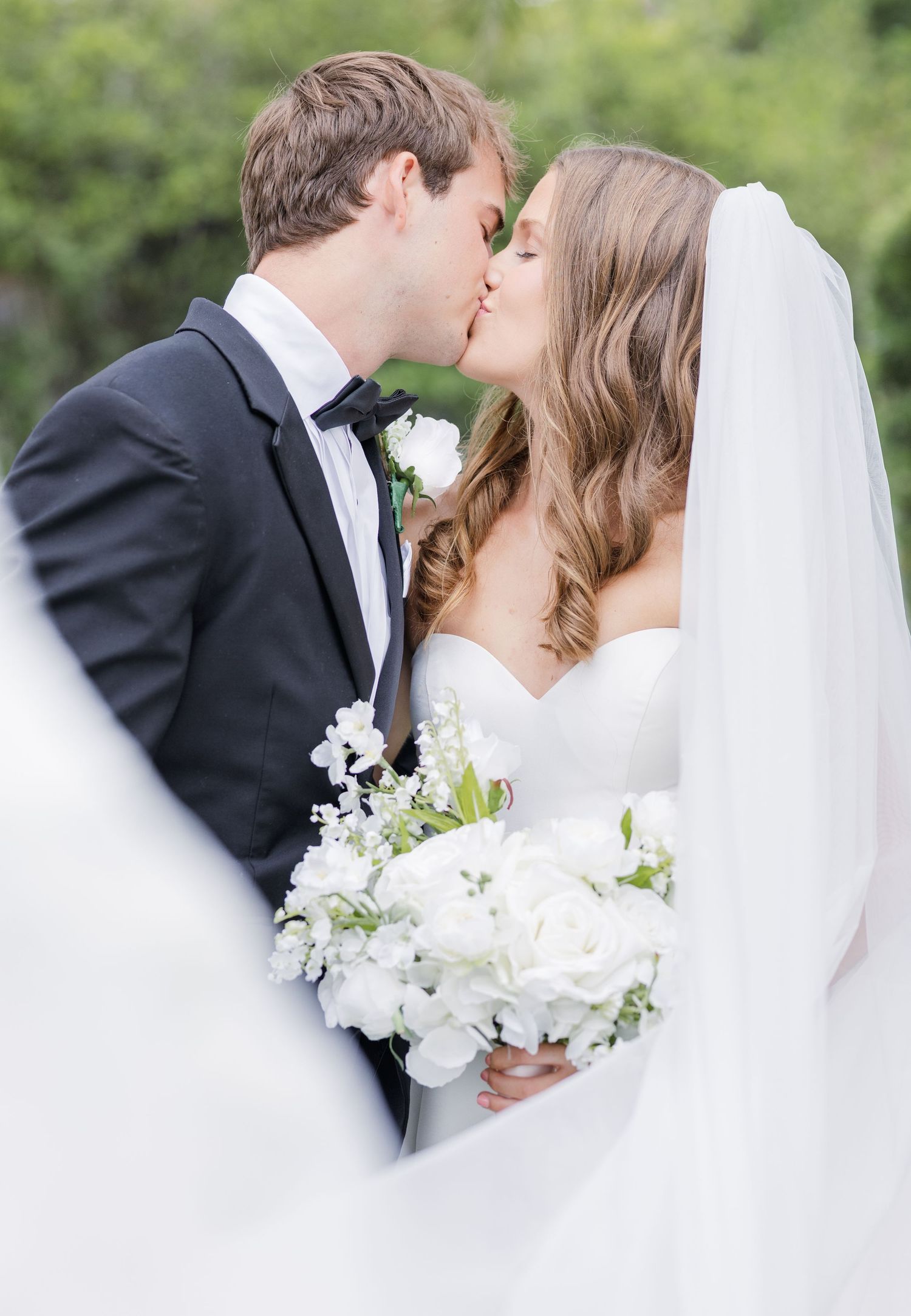 Bride and groom share a romantic kiss, bride holding white floral bouquet, groom in black tuxedo, outdoor wedding.