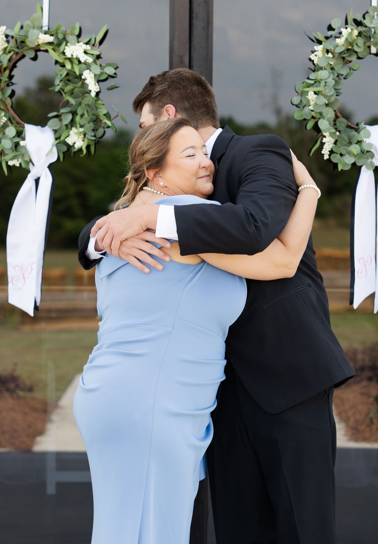Groom in black suit hugging woman in light blue dress at wedding ceremony with floral arch decorations.