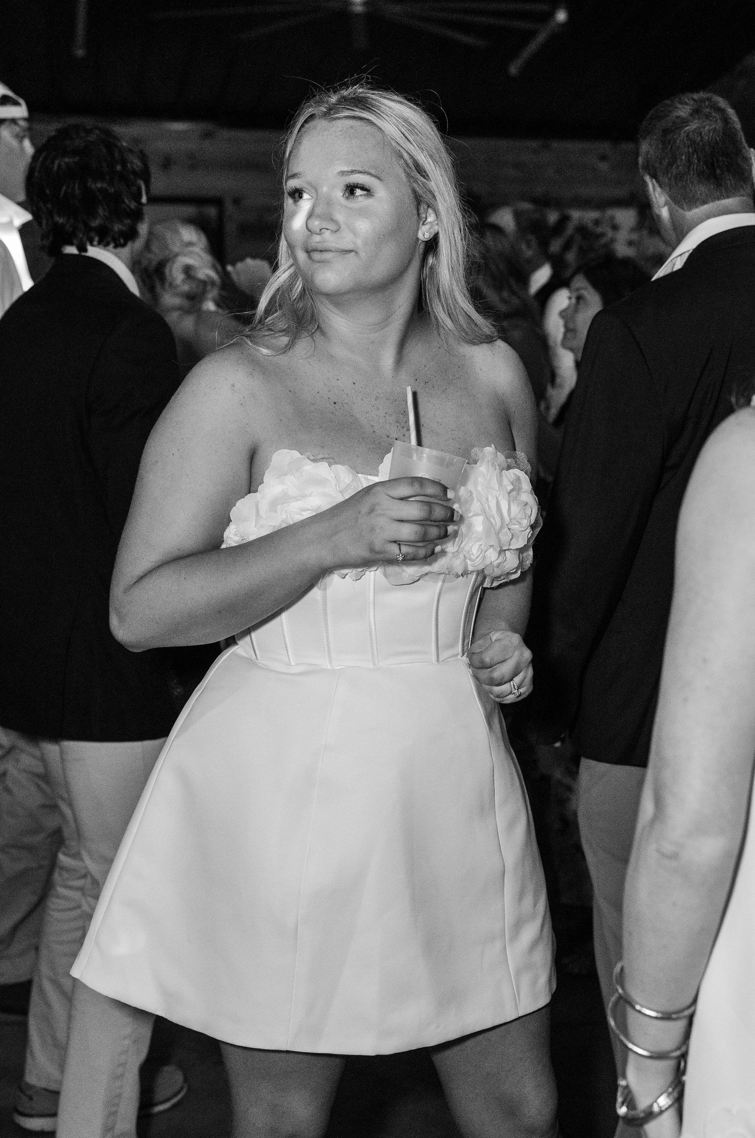 Young blonde woman in white strapless mini dress holds a drink at a wedding reception party.