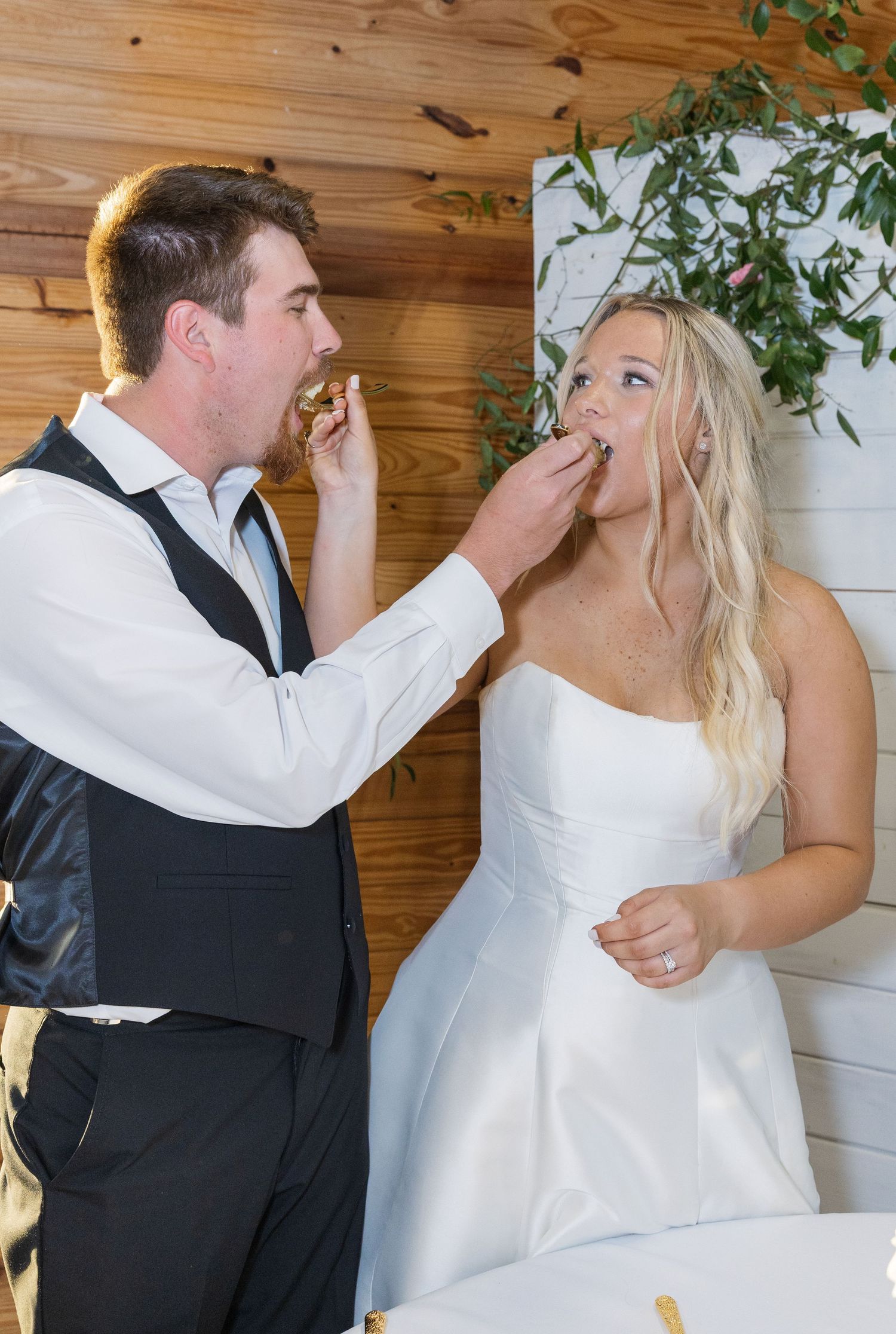Bride and groom feed each other wedding cake in front of rustic wood-paneled barn wall backdrop.