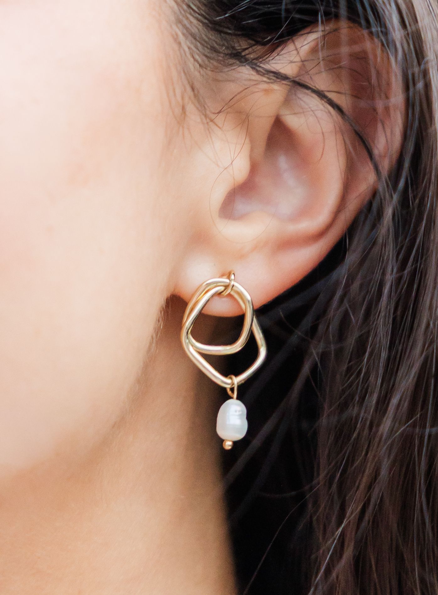 Close-up of gold double-loop earring with baroque pearl drop on a woman with dark hair.