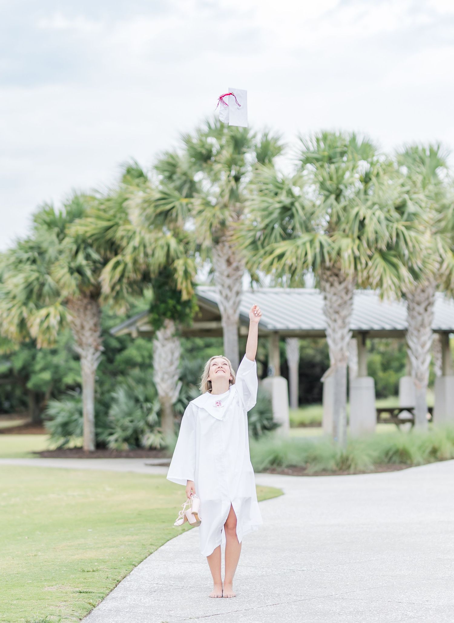 Graduate in white gown throws cap in celebration among palm trees.