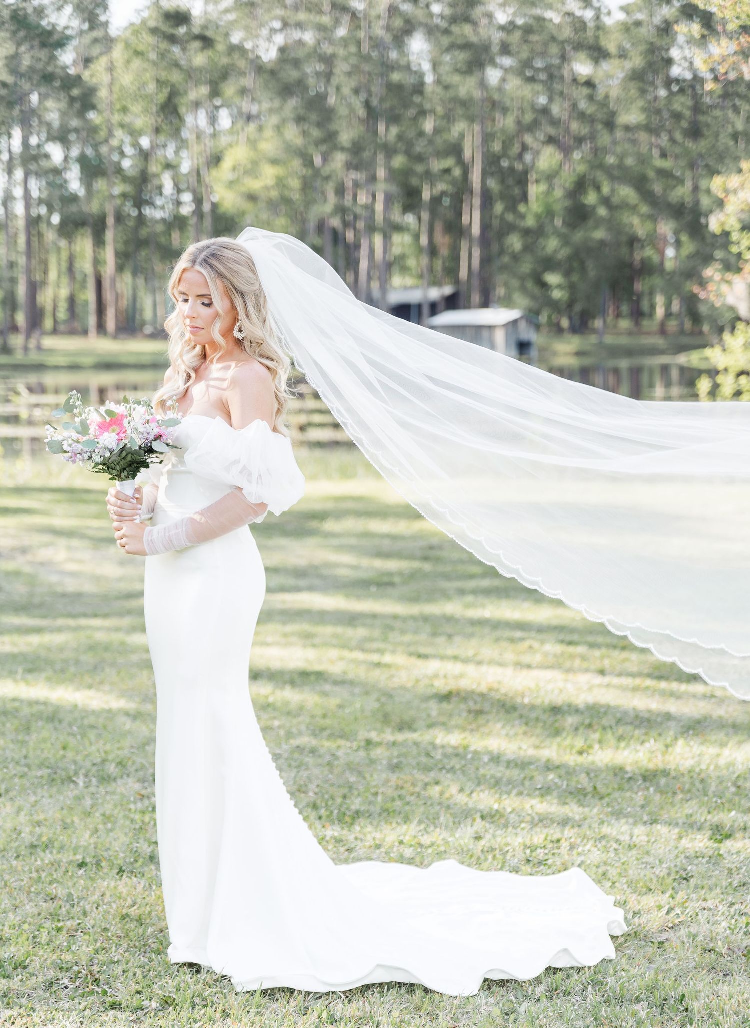 Bride with flowing cathedral veil holds bouquet while standing in sunlit garden setting.
