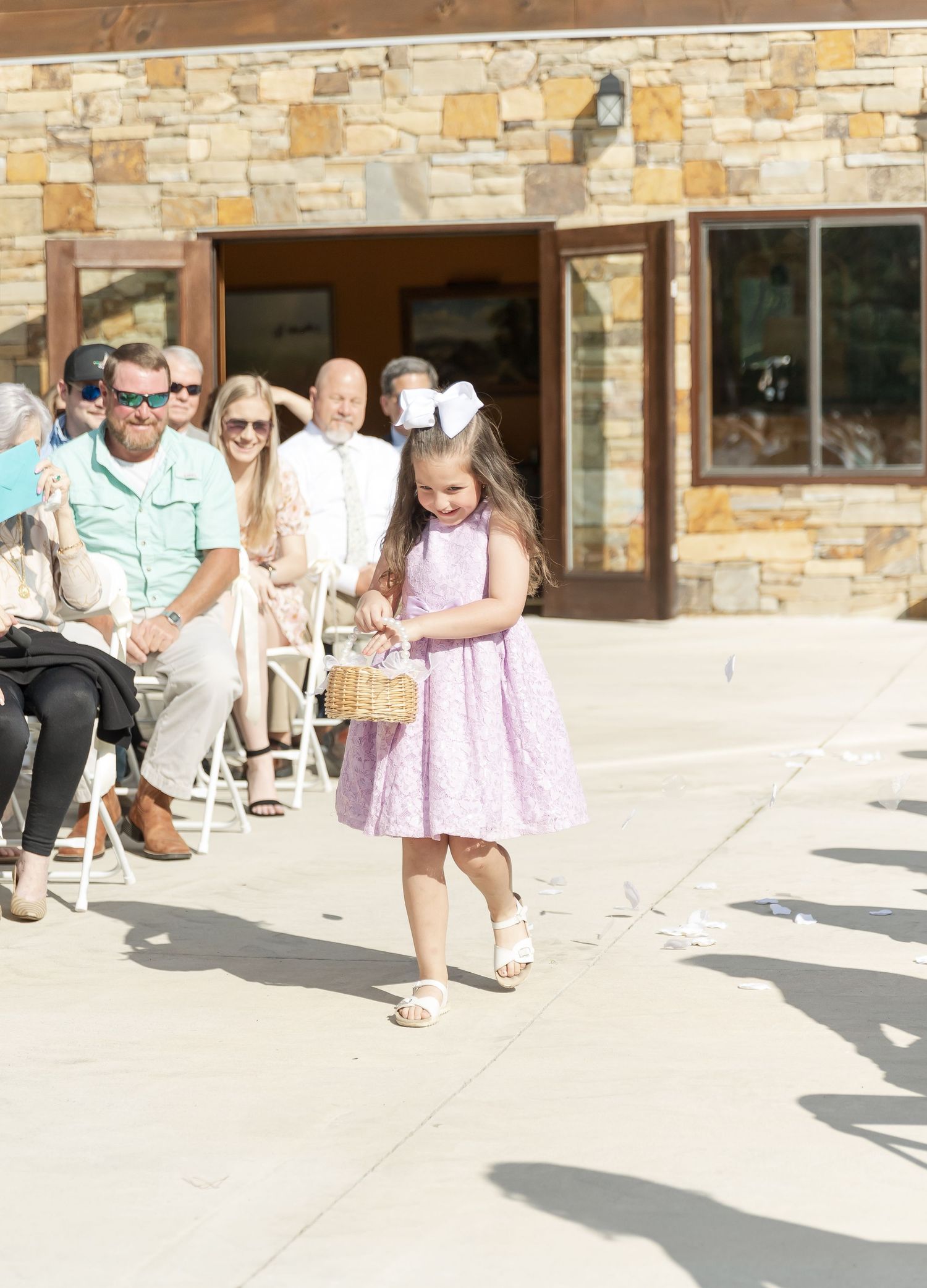 Young flower girl in pink dress walking down pathway at outdoor venue with stone walls.
