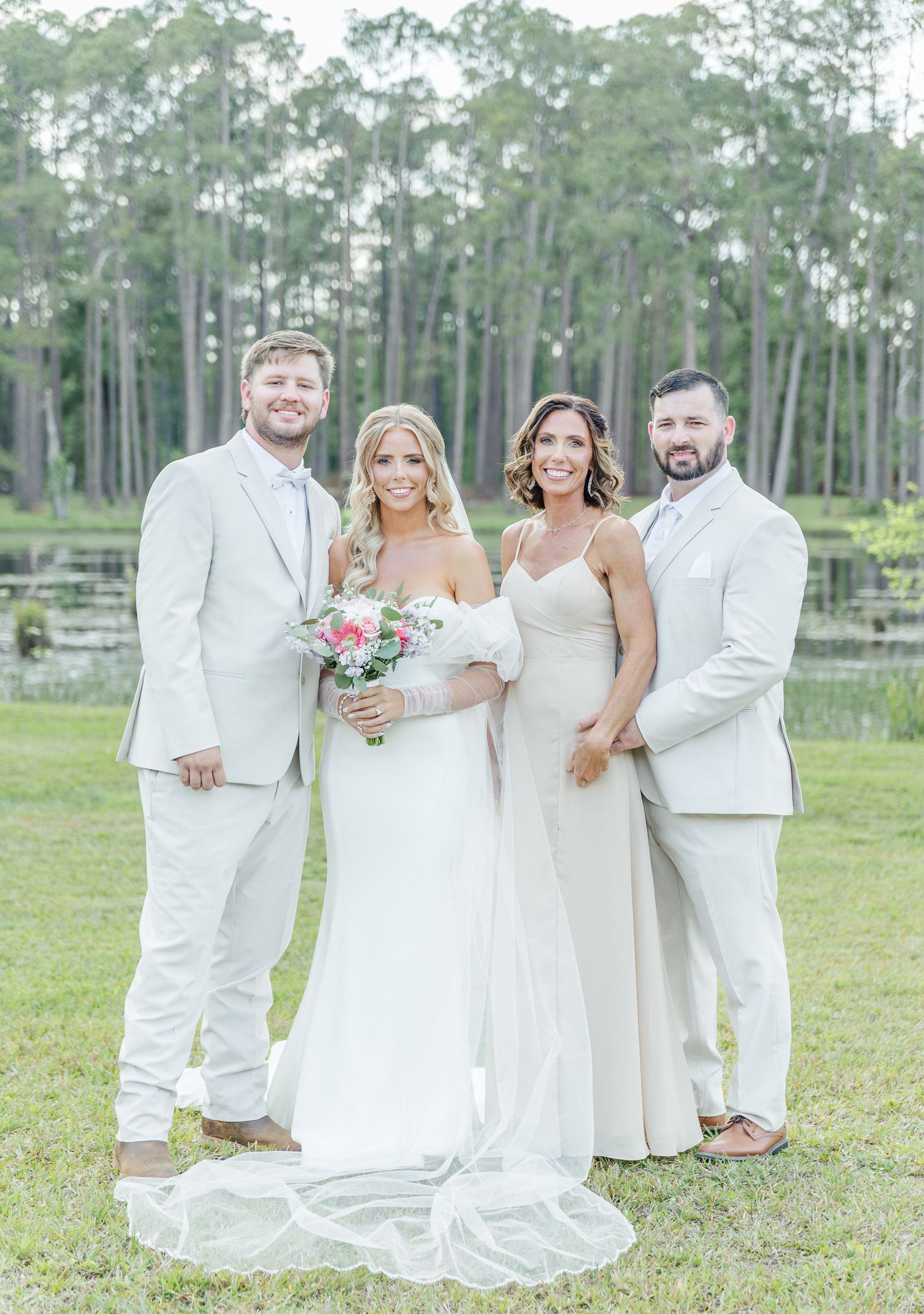 Two couples in formal white attire pose for wedding photos in front of a scenic forest landscape.