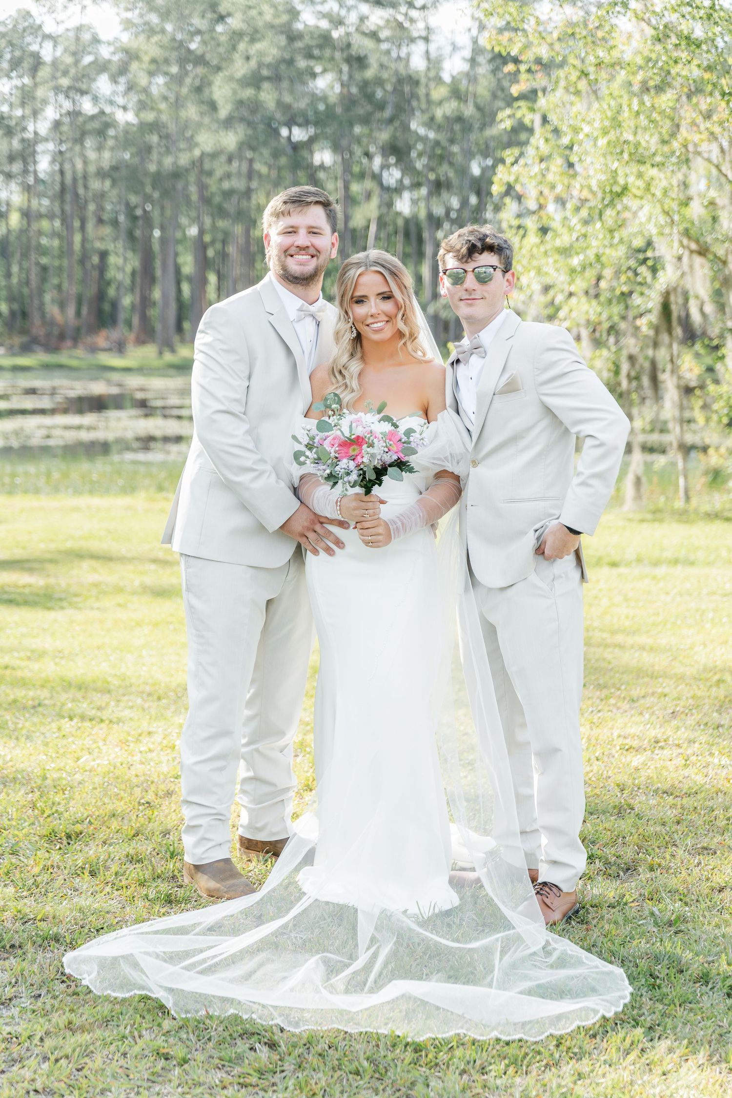 Three people in white attire pose with bouquet in outdoor wedding setting.