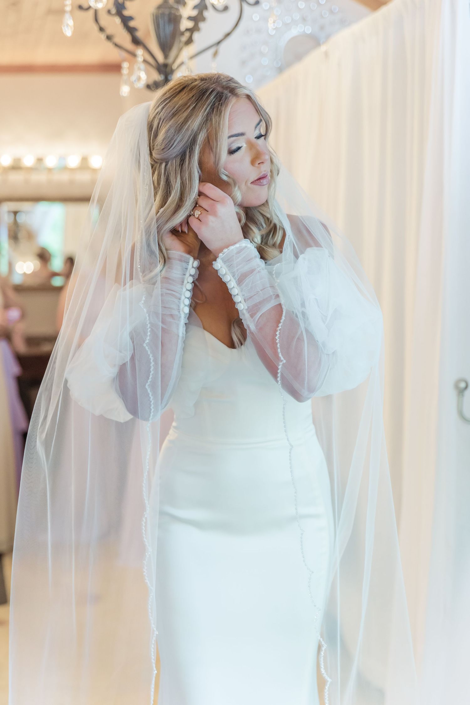 A bride in a fitted white wedding dress with sheer sleeves adjusts her long veil in an elegant indoor setting.