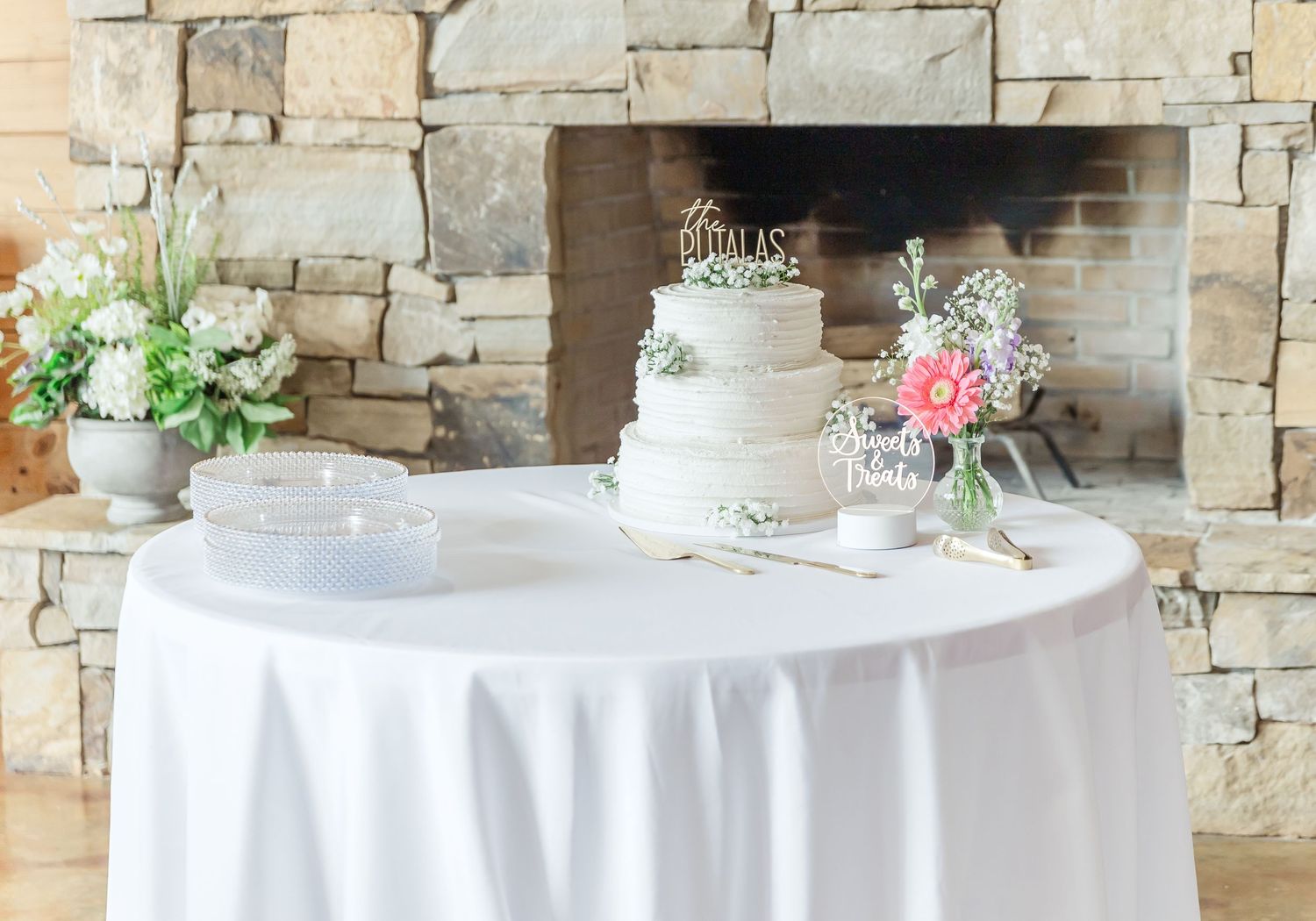 Three-tier white wedding cake displayed on table with stone fireplace backdrop.