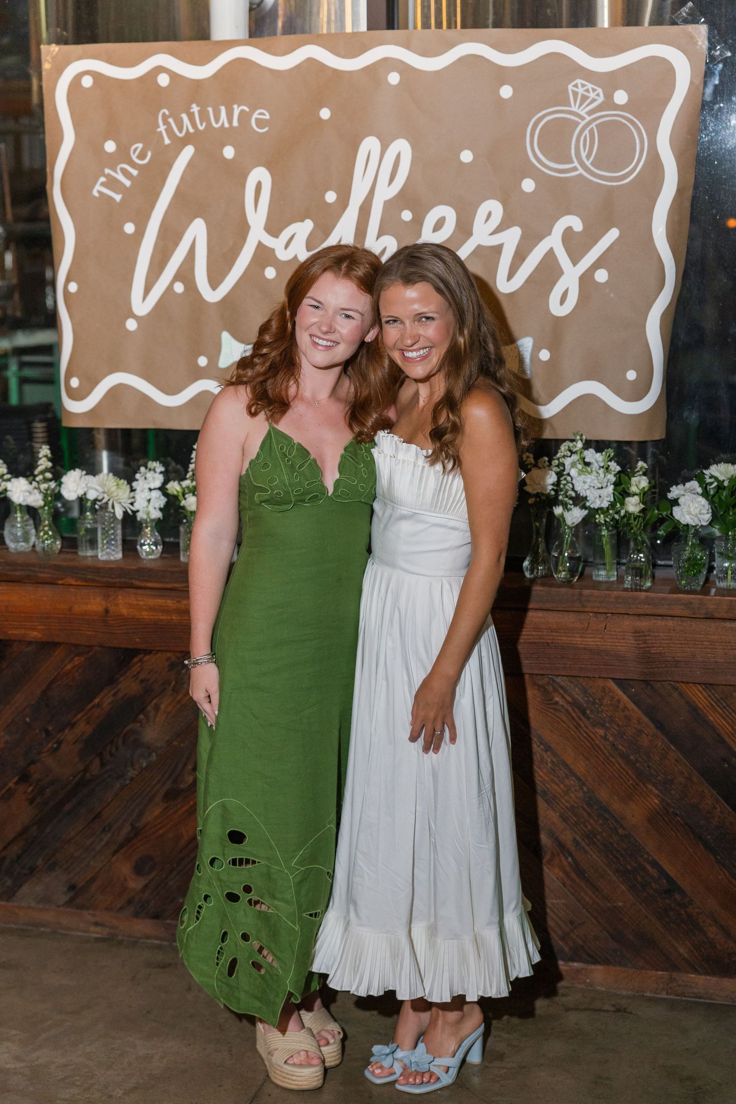 Two women smile at engagement party before 'The Future Walkers' sign, one in green, one in white.