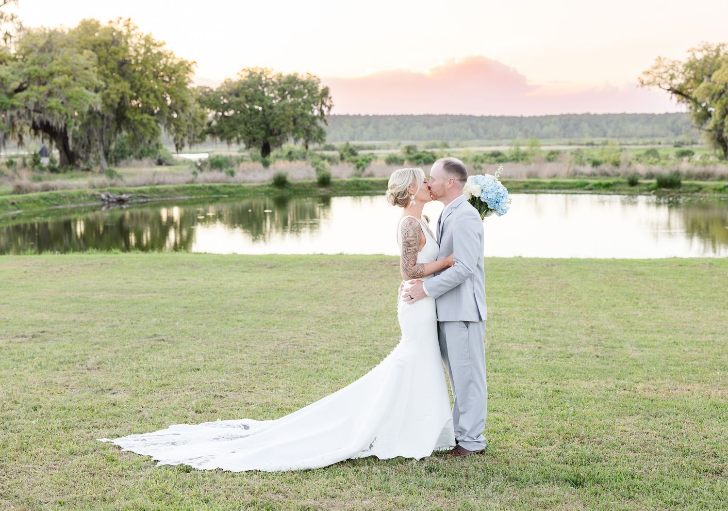 Bride and groom kissing by a pond at sunset with mountain backdrop.