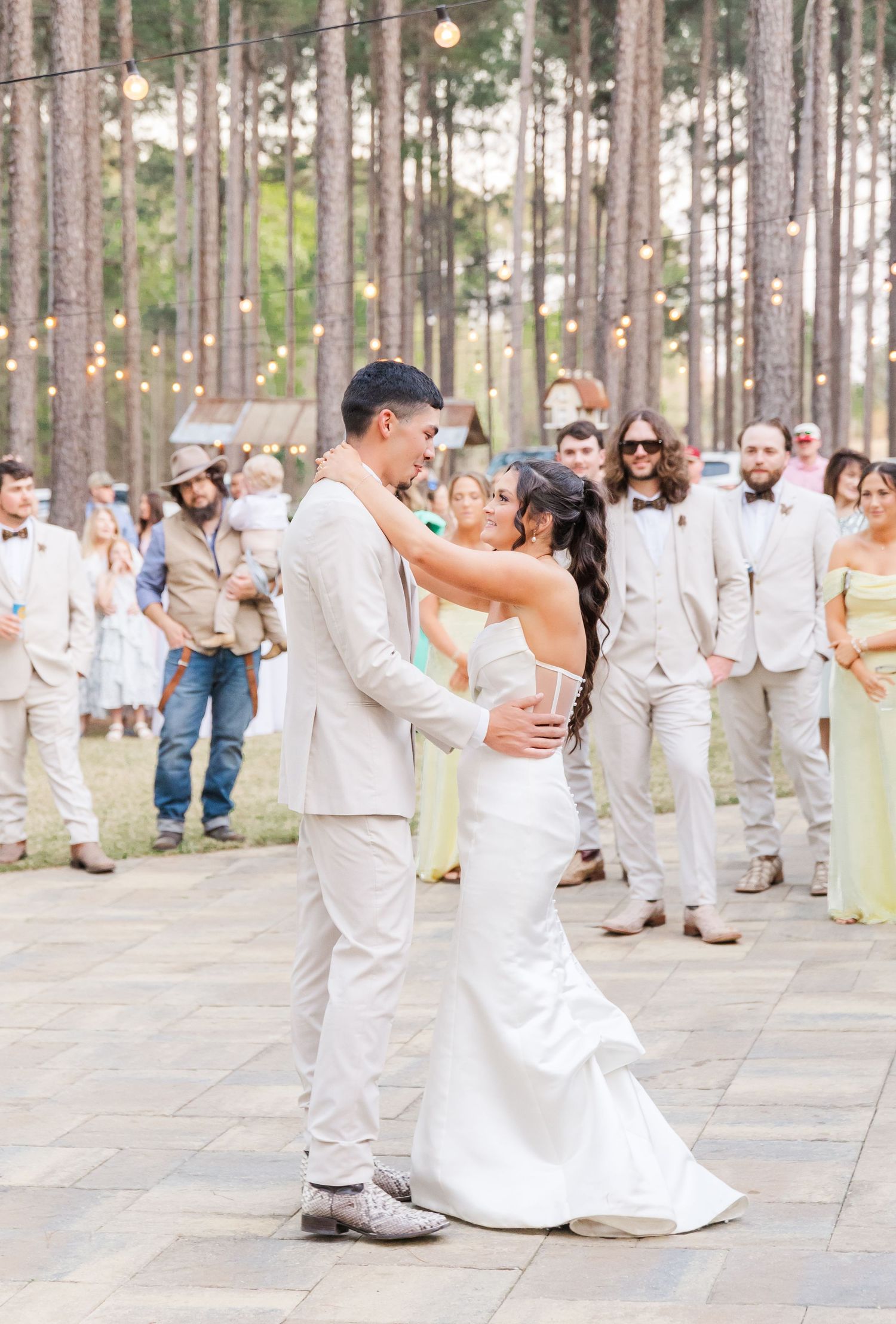 Bride and groom share first dance on outdoor patio surrounded by guests and string lights at reception.