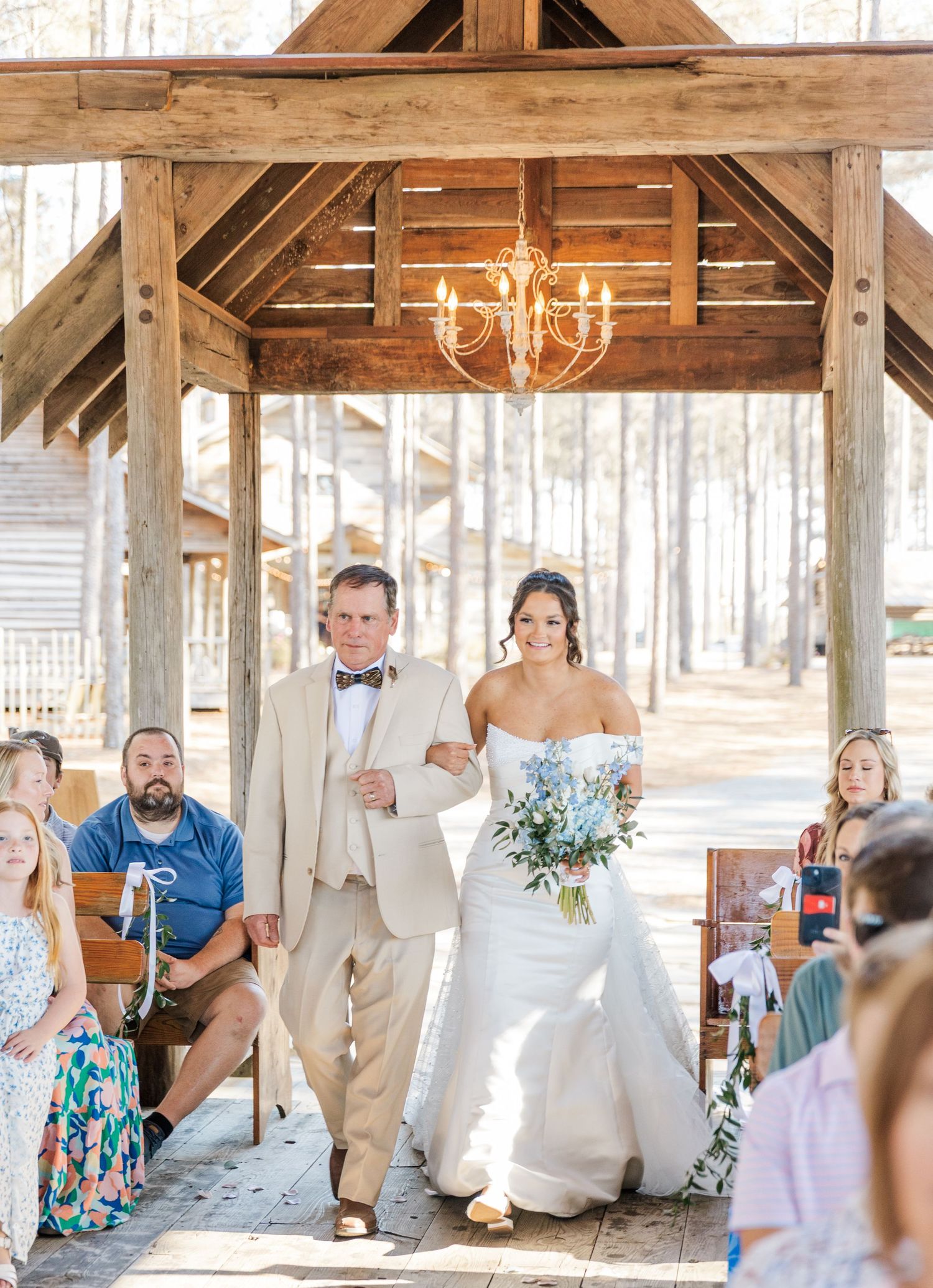 Bride walks down outdoor aisle under rustic wooden gazebo with chandelier escorted by her father.
