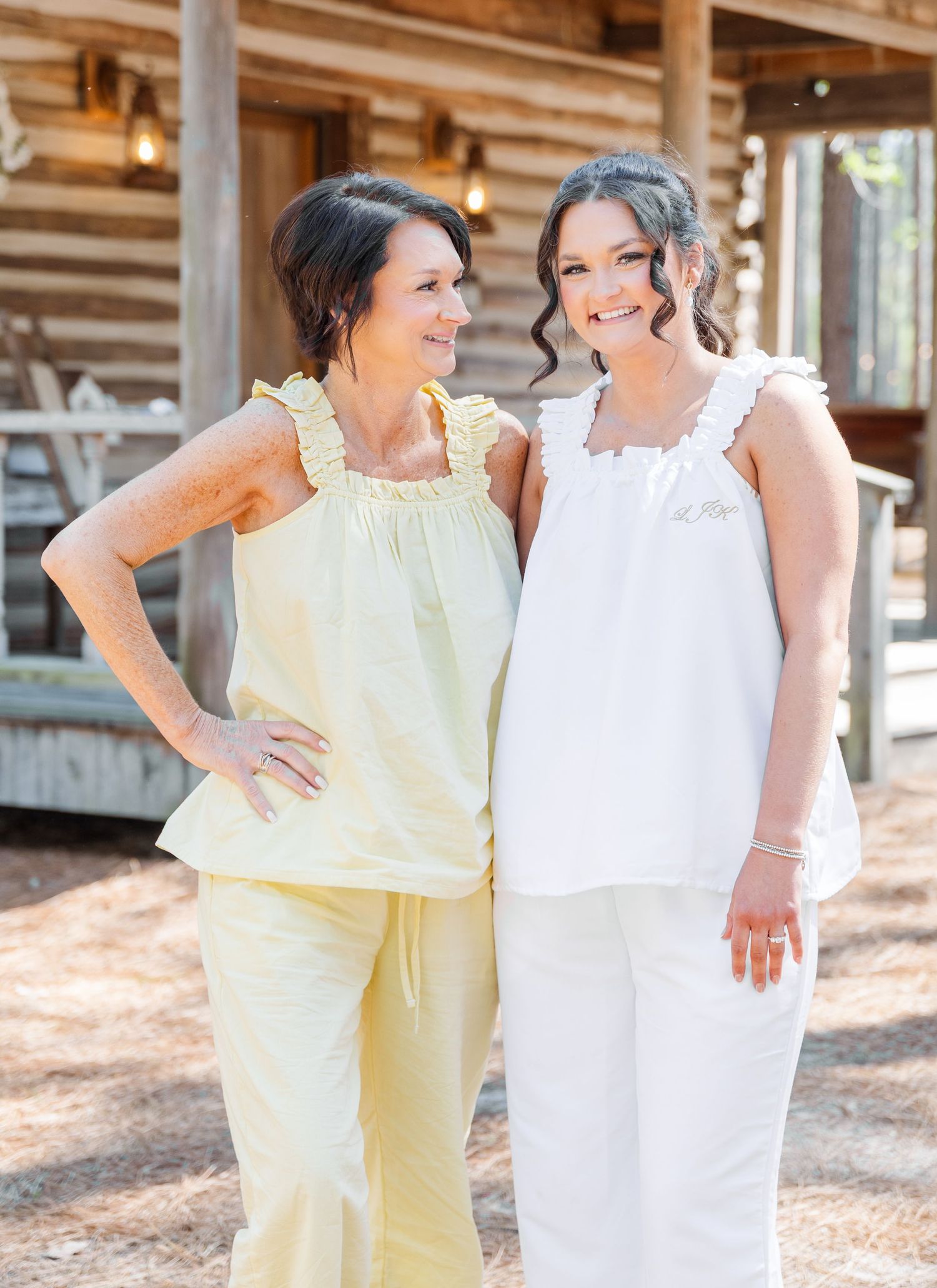 Mother and bride smiling together in matching yellow and white ruffled outfits outside a log cabin.