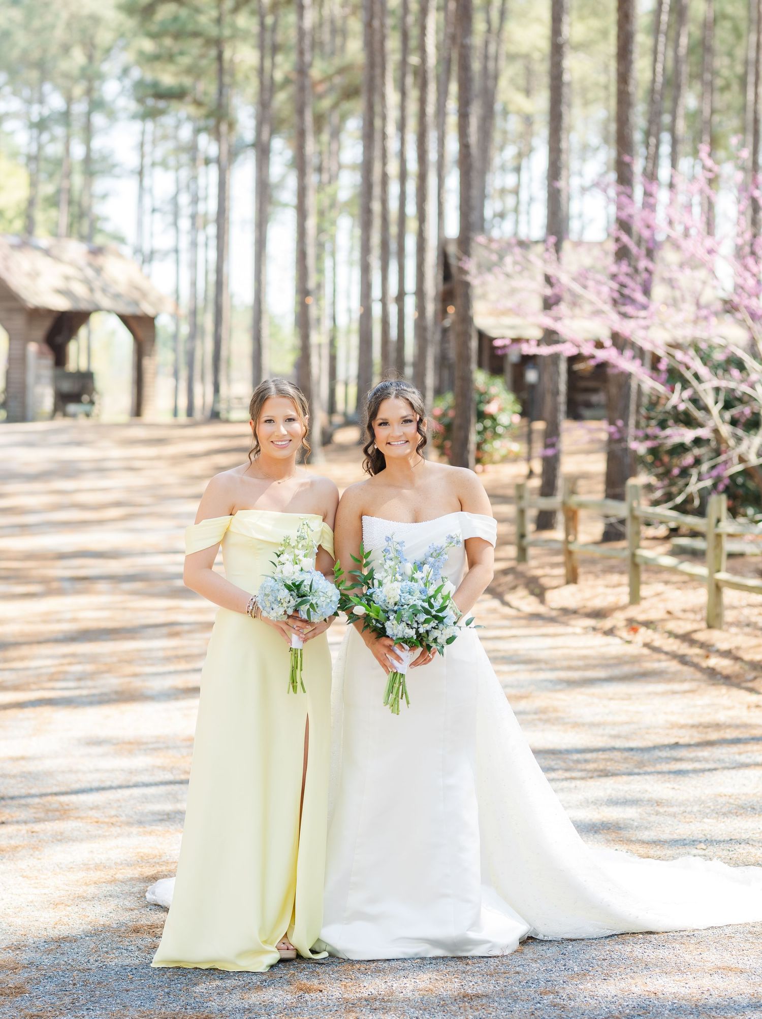 Bride in off-shoulder white gown poses with bridesmaid in yellow dress holding blue bouquets.