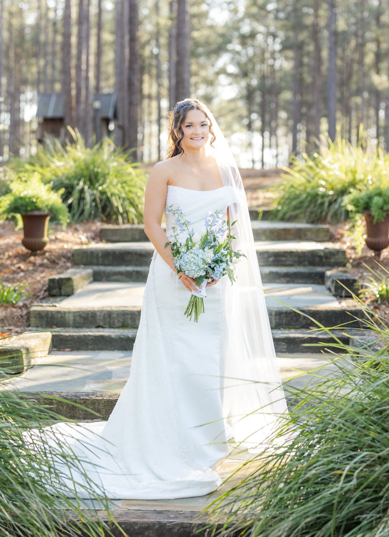 Smiling bride in strapless white gown holds blue bouquet on garden steps surrounded by pines.