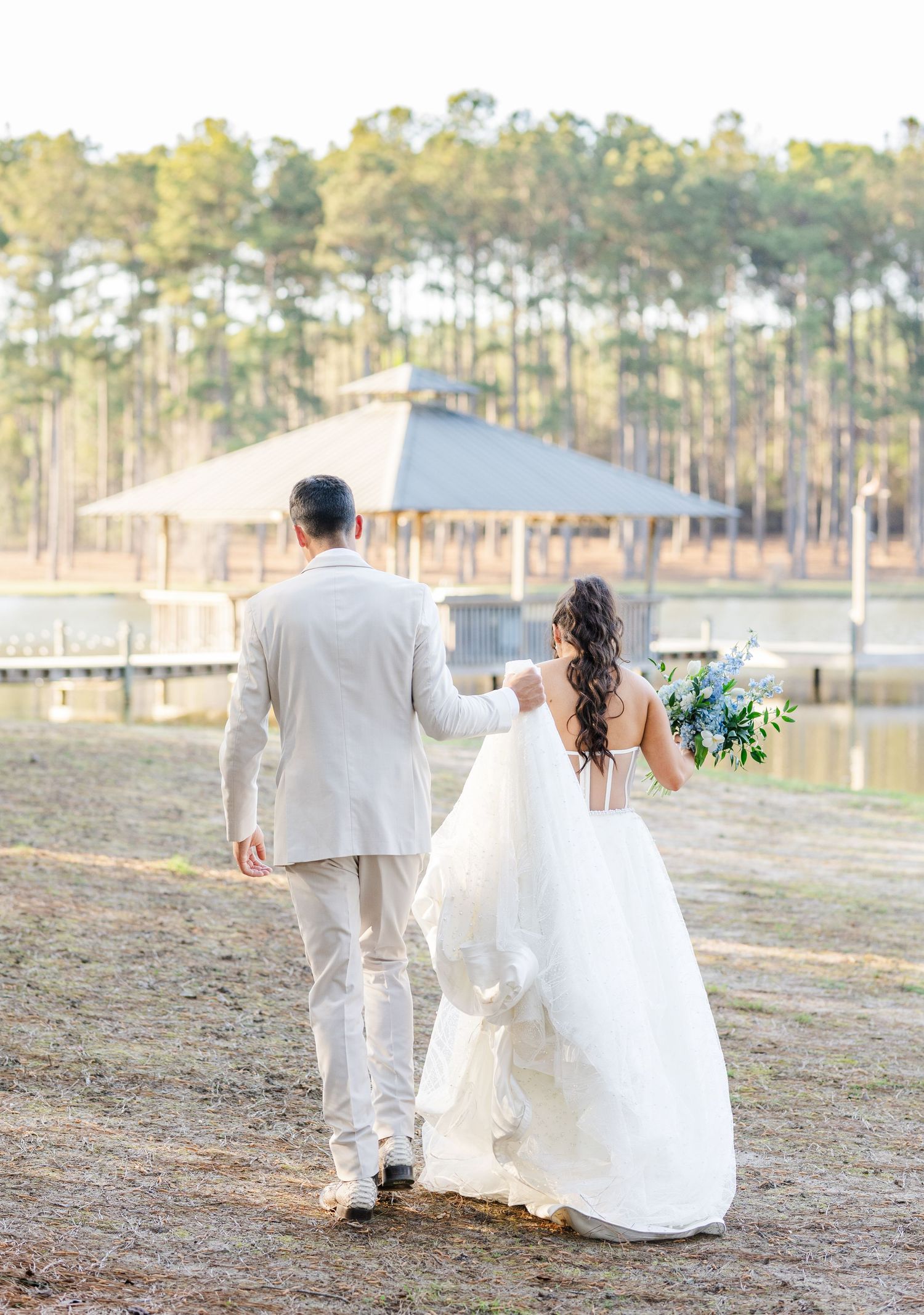 Bride and groom walking hand in hand toward a lakeside gazebo, bride holding a blue and white bouquet in a white ballgown.