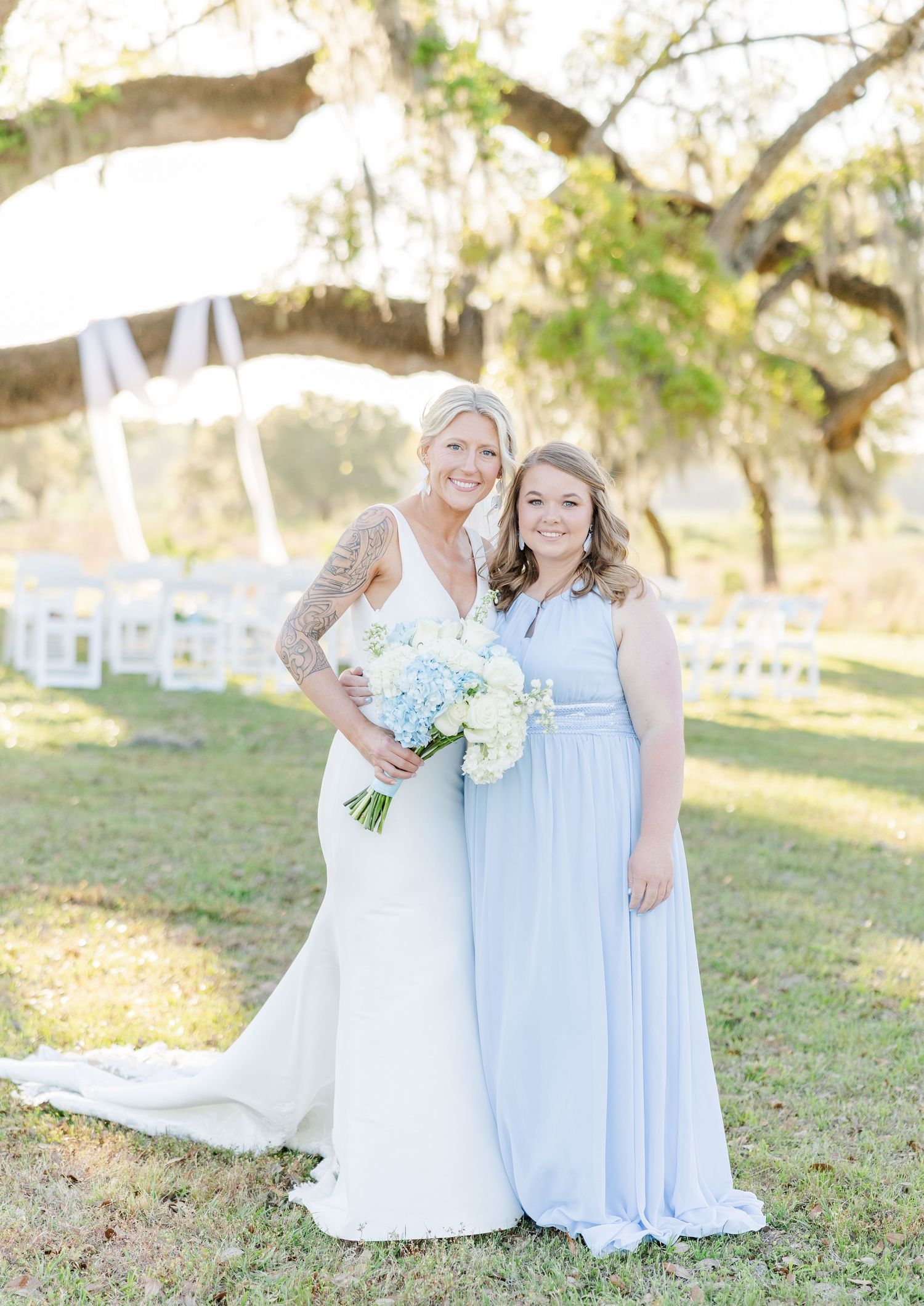 Bride in white gown holding blue and white bouquet poses with bridesmaid in light blue dress under oak trees.