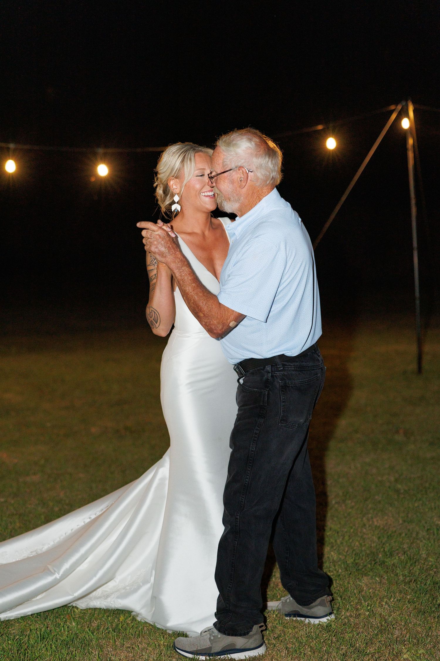 Bride in white satin gown dancing with elderly man under string lights at nighttime outdoor reception.