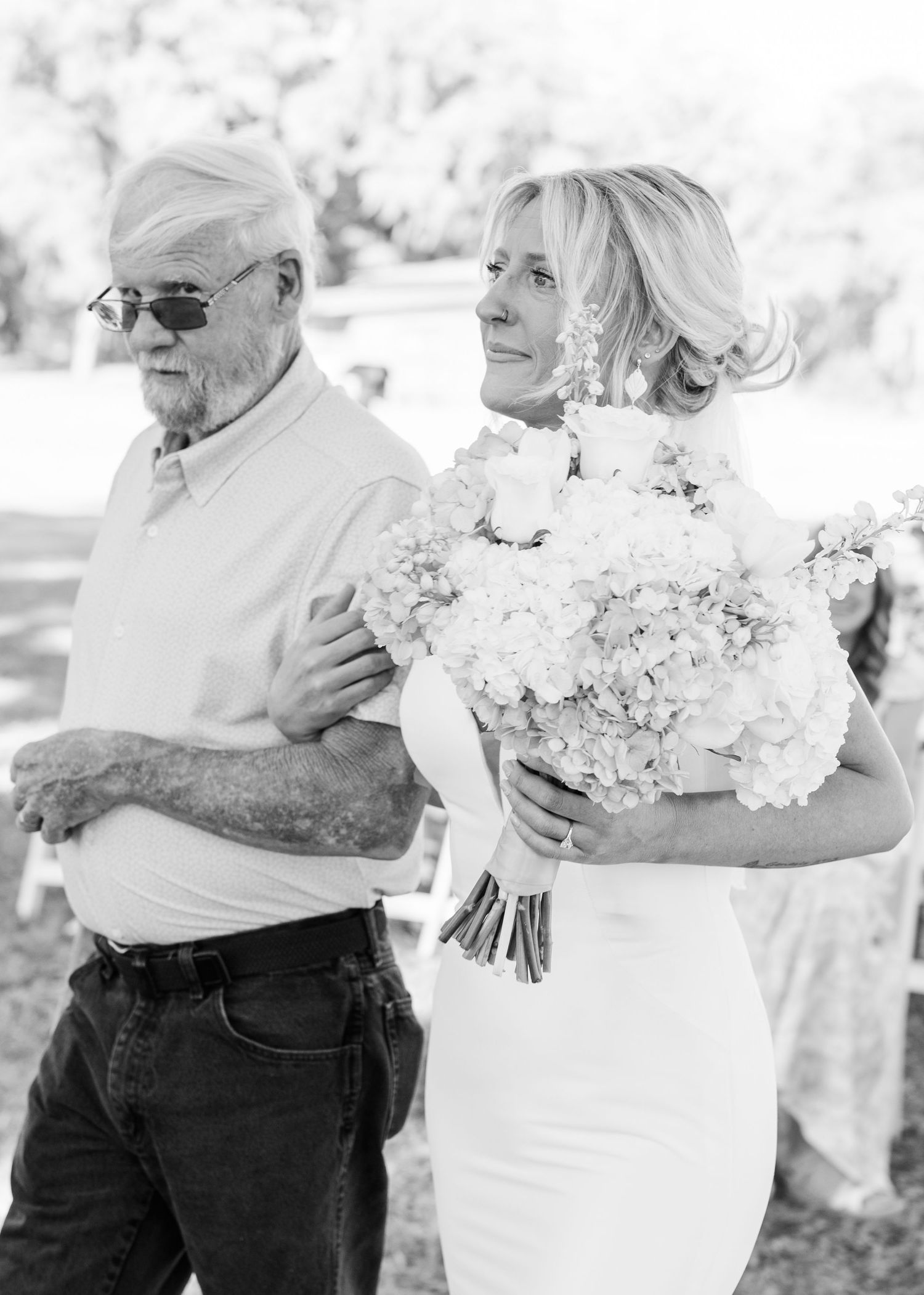Black and white photo of bride holding bouquet walking down aisle with older gentleman escort at outdoor wedding.