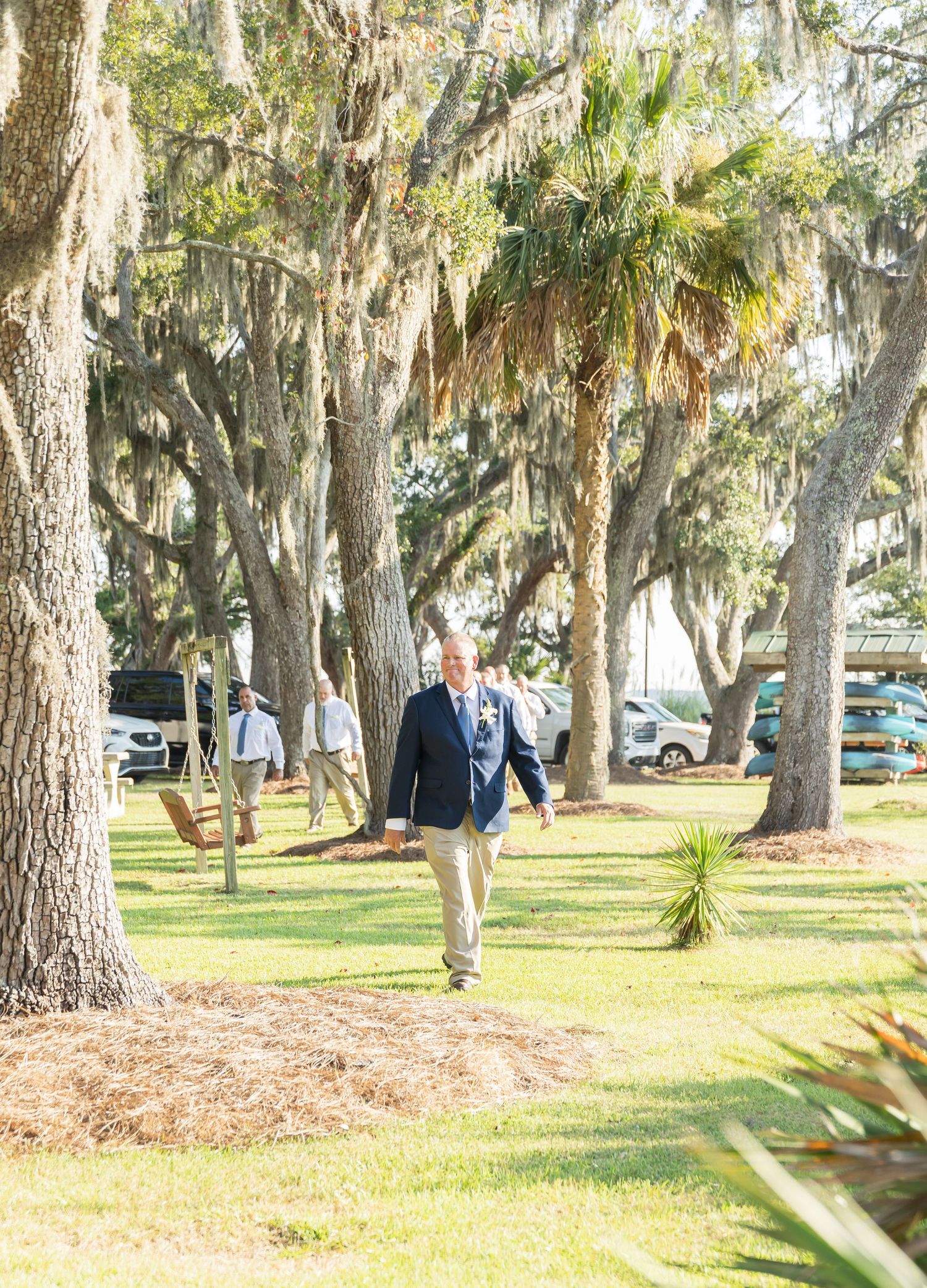 Spanish moss hangs from oak trees in a scenic park setting with parked cars visible in the background.