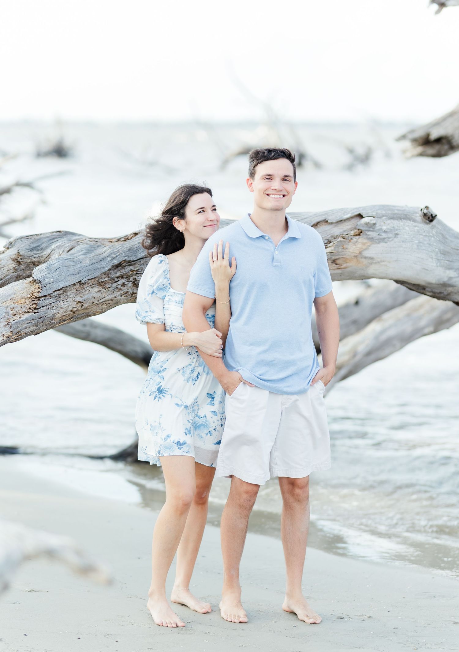Couple strolls barefoot along the ocean's edge near weathered driftwood on a bright beach.