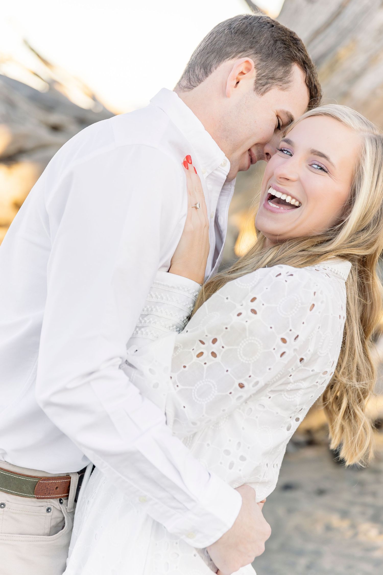 A romantic beach photoshoot showing a couple in white outfits laughing and embracing in golden sunlight.