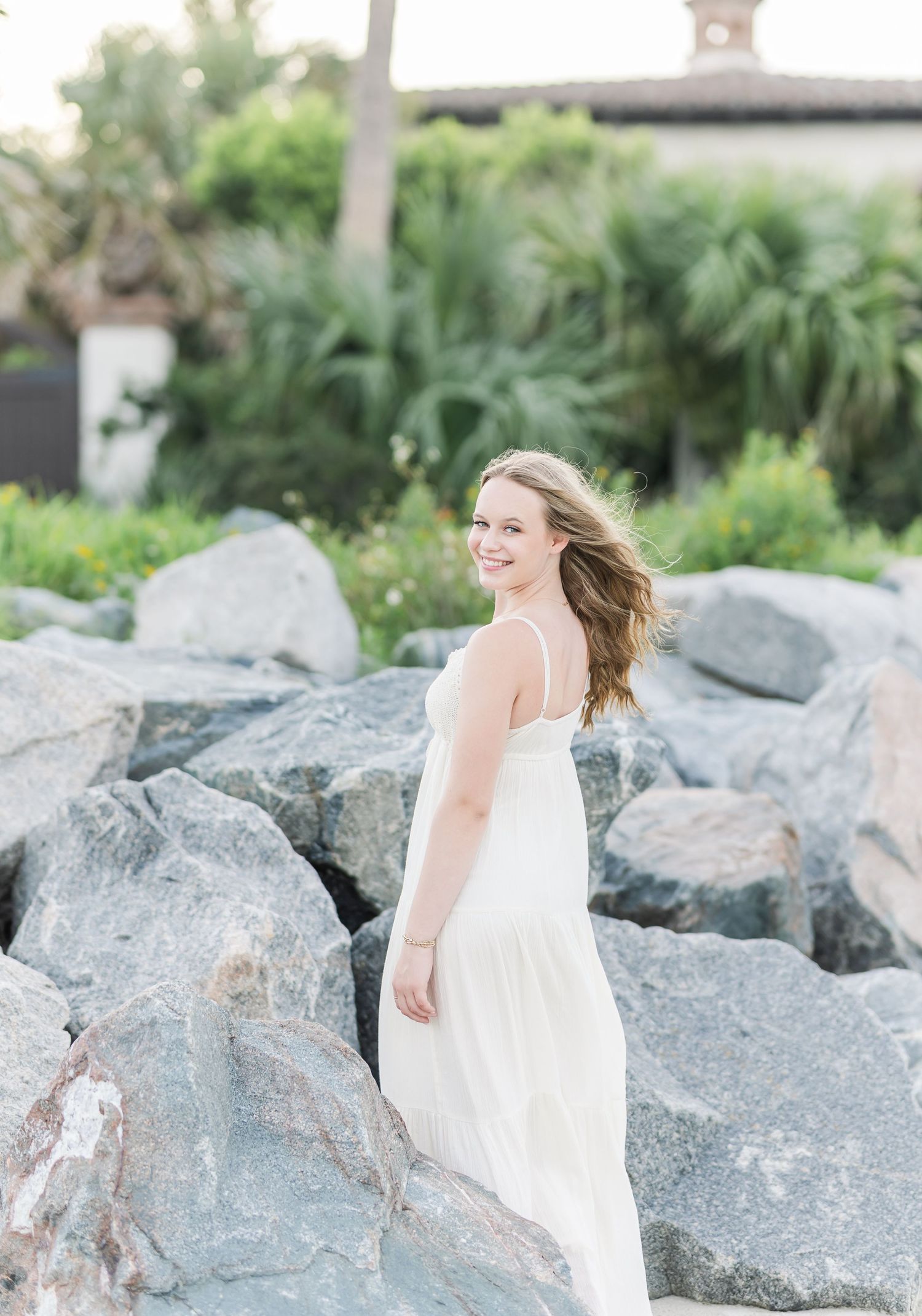 Person in white sundress moves joyfully among large coastal rocks in a lush garden setting.