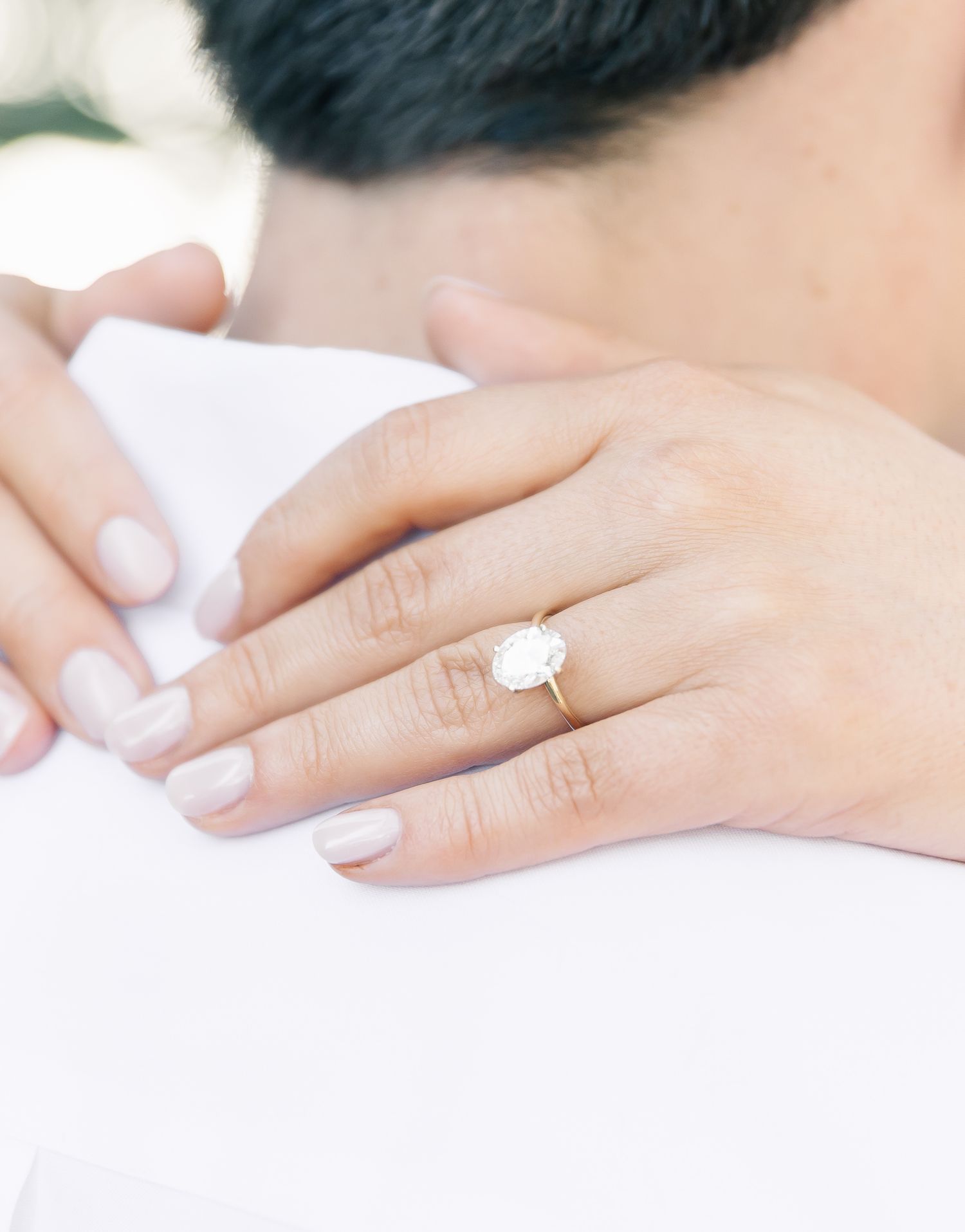 Close-up shot of hands wearing a delicate oval engagement ring with white manicured nails.
