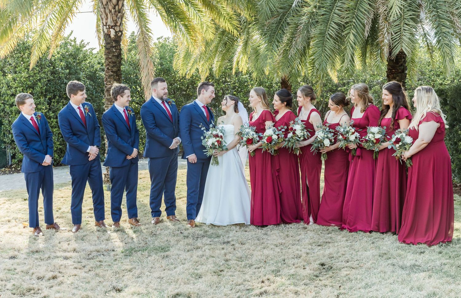 Wedding party in navy suits and burgundy dresses poses together under palm trees.