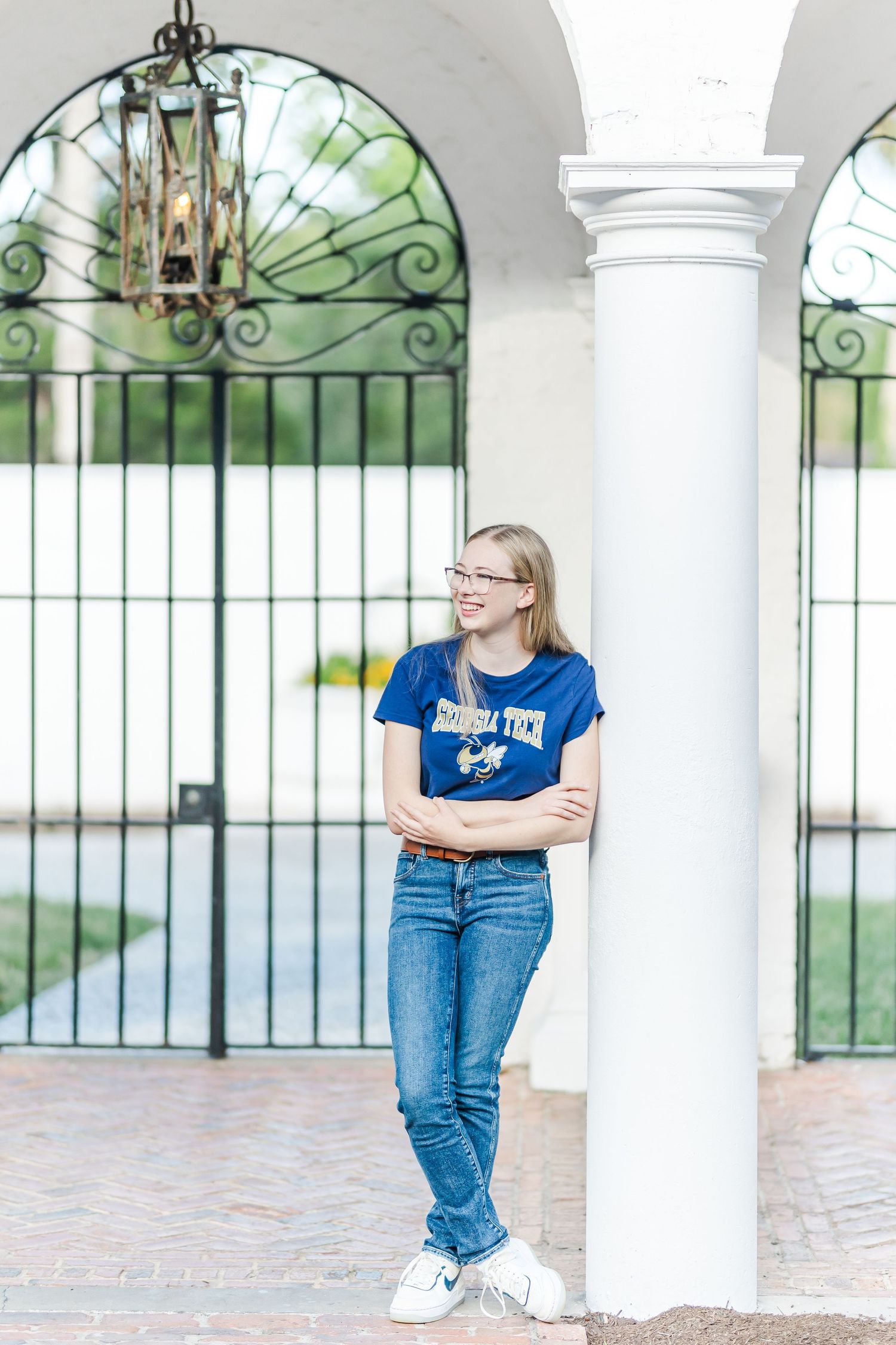 Person in blue university shirt poses between ornate iron gates and white columns.