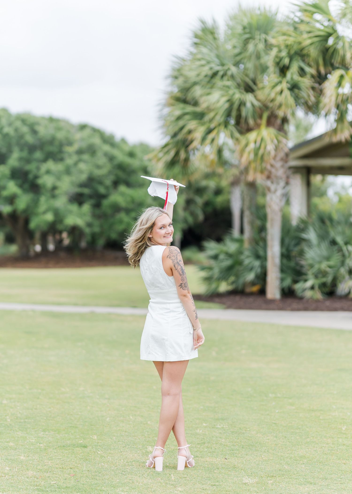 Graduate poses on green lawn holding cap overhead with palm trees in background.