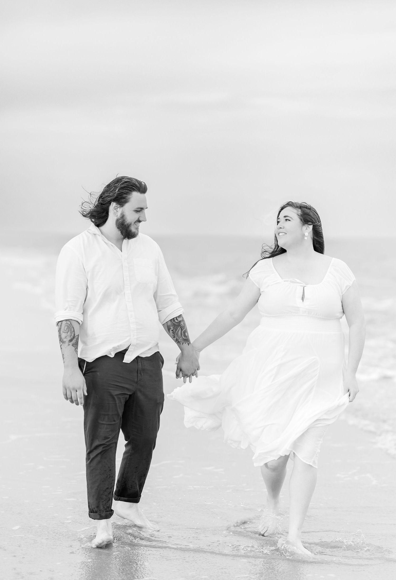 A couple walks hand in hand along the beach shore in black and white photography.