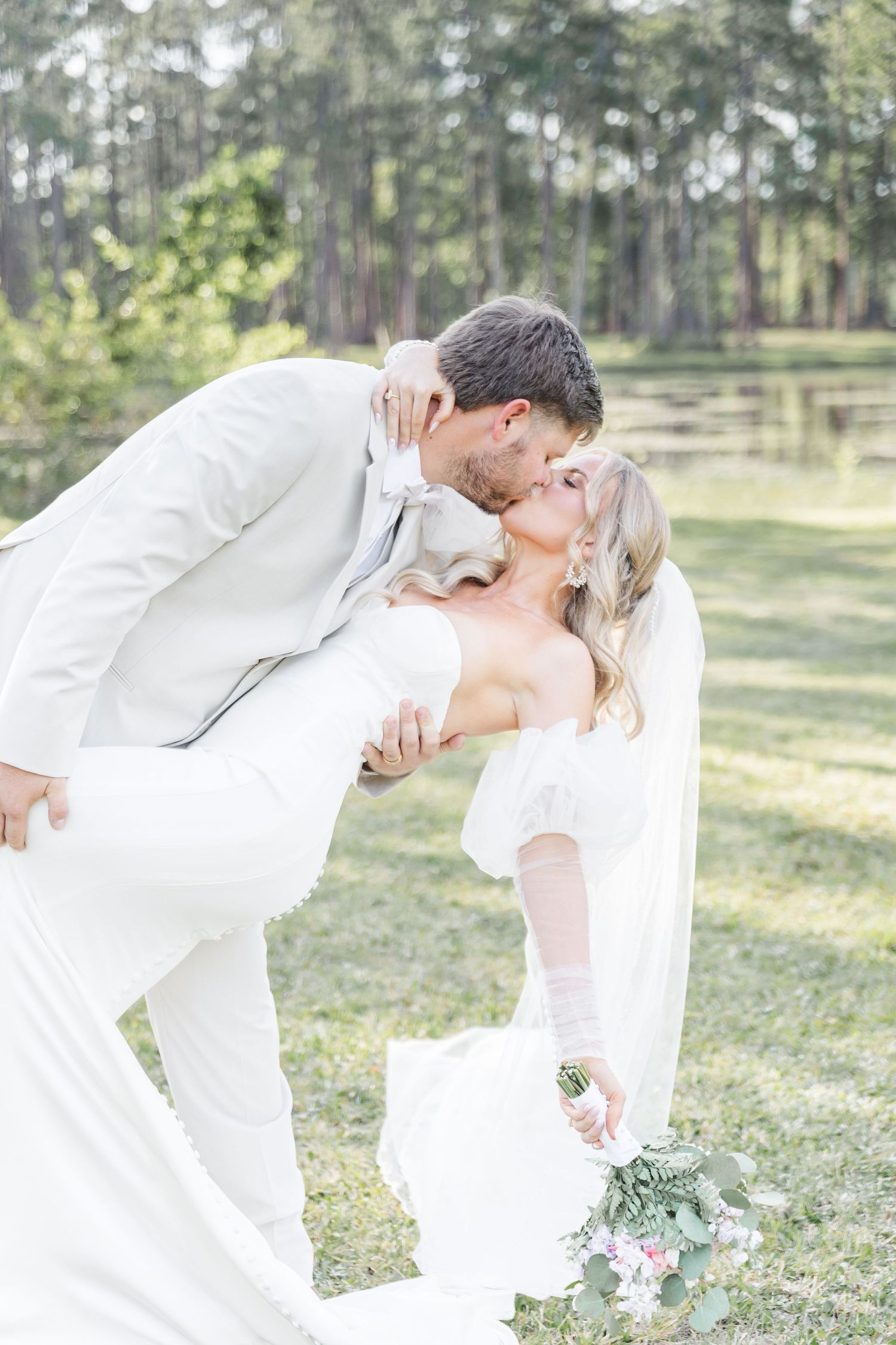 A romantic dip kiss between newlyweds wearing all white against a backdrop of tall pine trees.