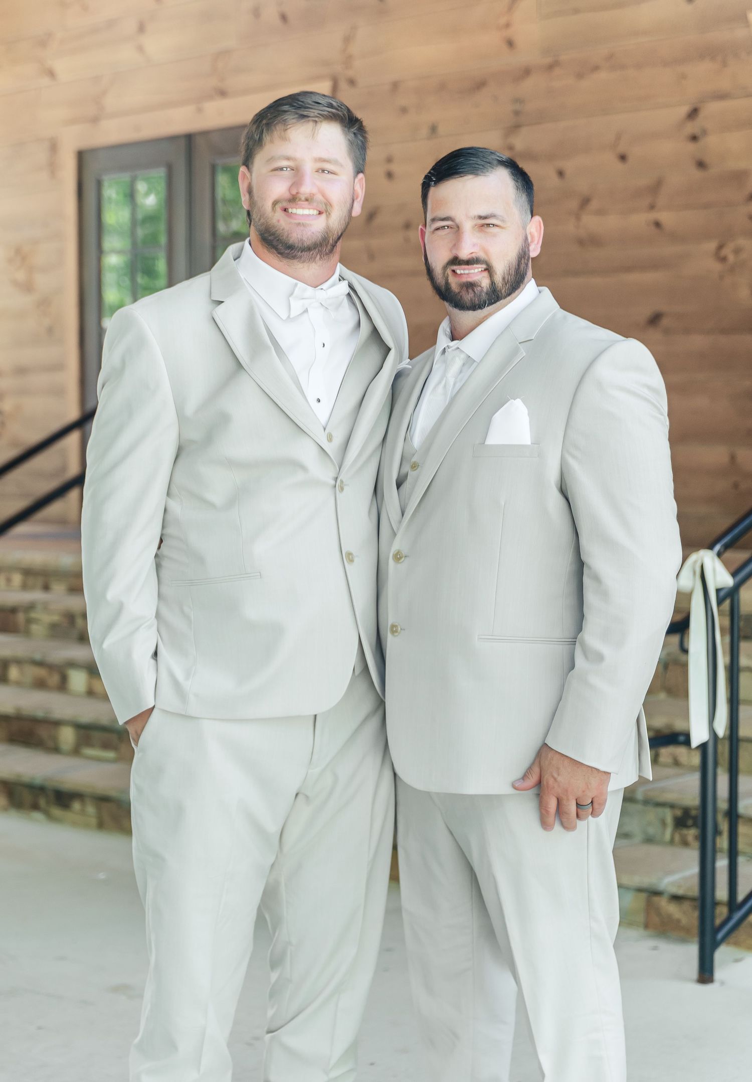 Two people wearing coordinated cream suits pose together outside a wooden building.