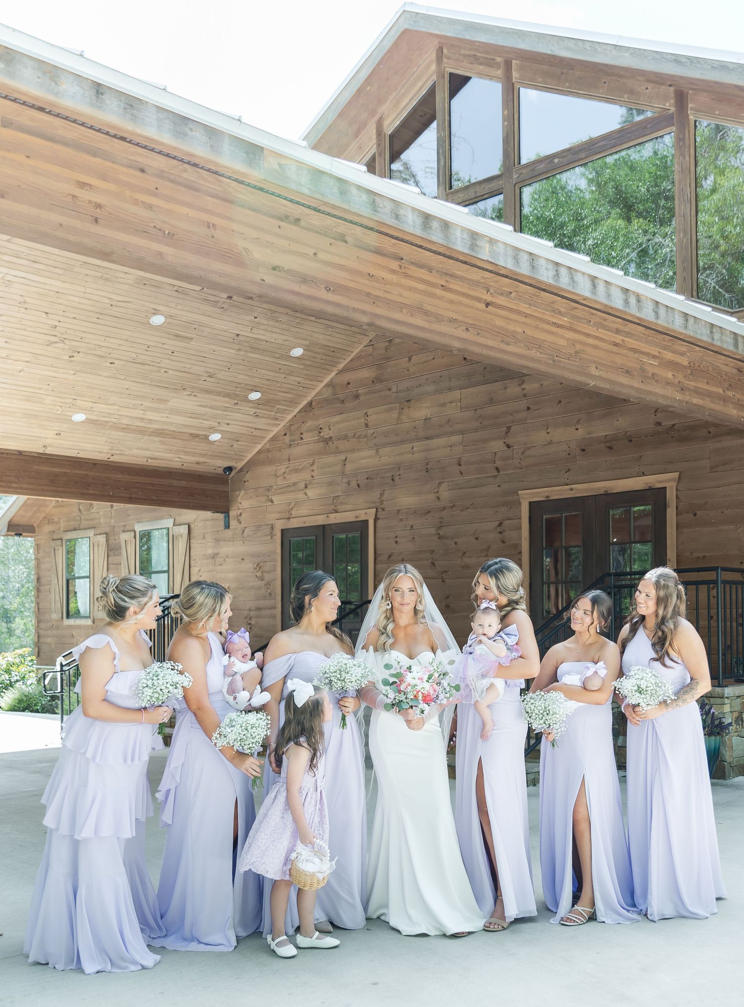Bridal party in pale lavender dresses pose with bouquets under rustic wooden porch overhang.