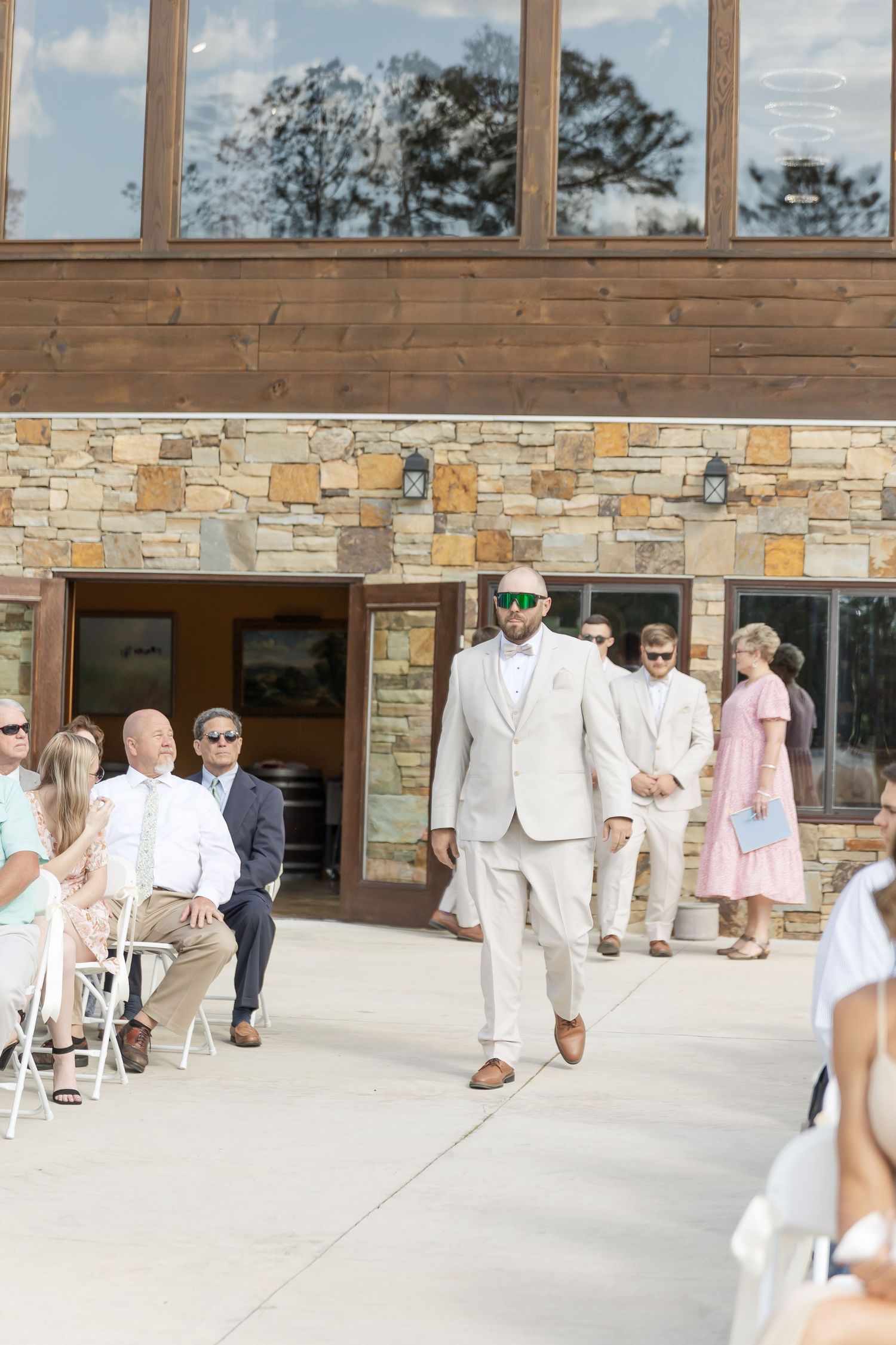 Wedding attendant in light beige suit and sunglasses walks down outdoor ceremony aisle.