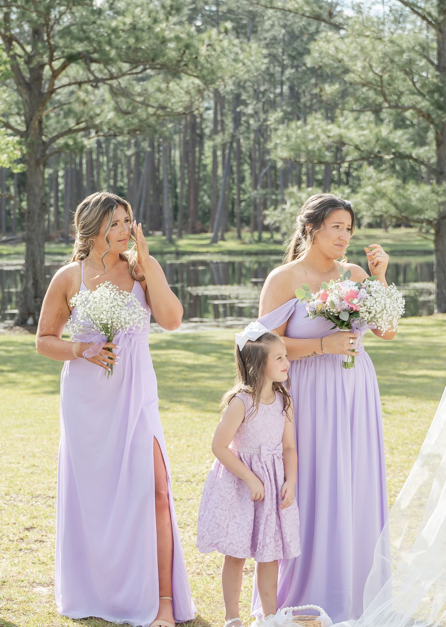 Wedding party members in lavender dresses gather outdoors near a serene pond with lush greenery.