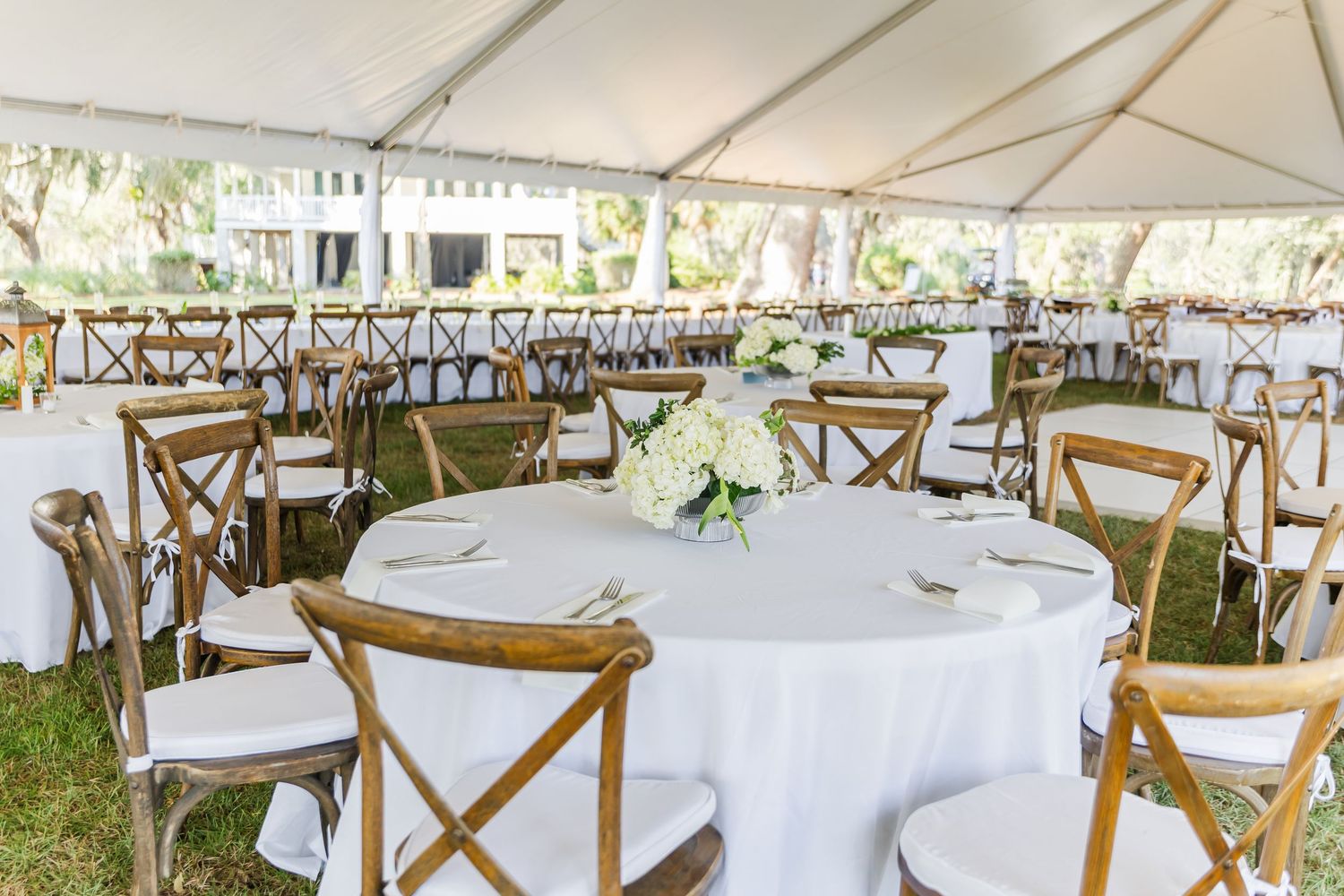 Elegant outdoor wedding reception setup with white tablecloths and wooden cross-back chairs under a white tent.
