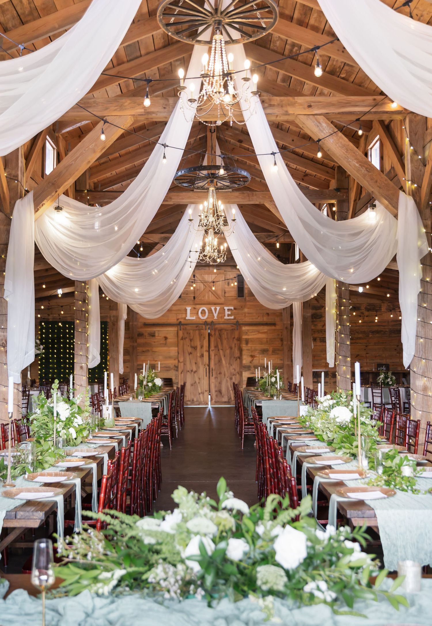 Rustic barn wedding reception venue decorated with white draping, chandeliers and long tables with greenery centerpieces.