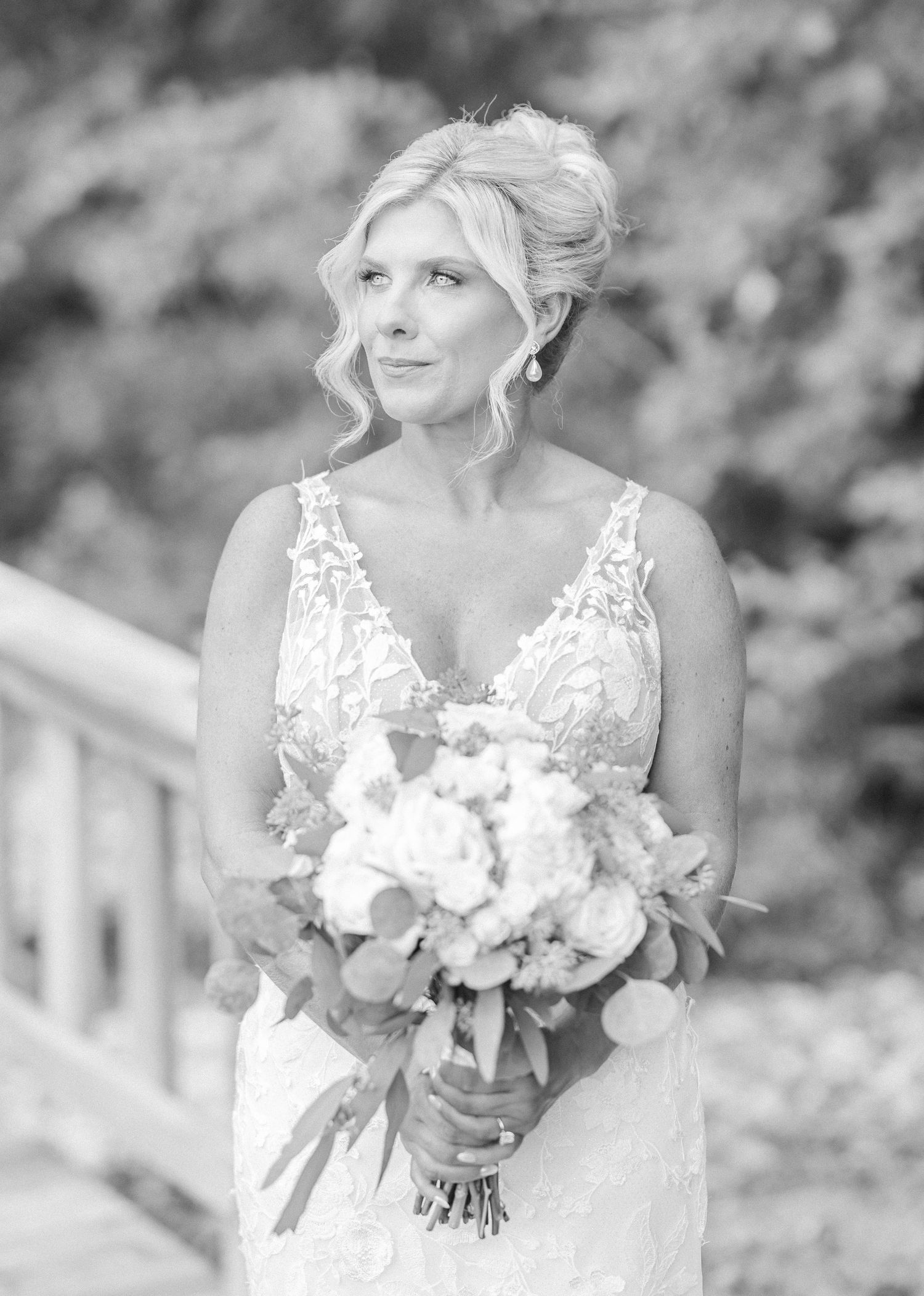 A bride in a white lace dress holds a beautiful bouquet of white flowers against a blurred outdoor background.