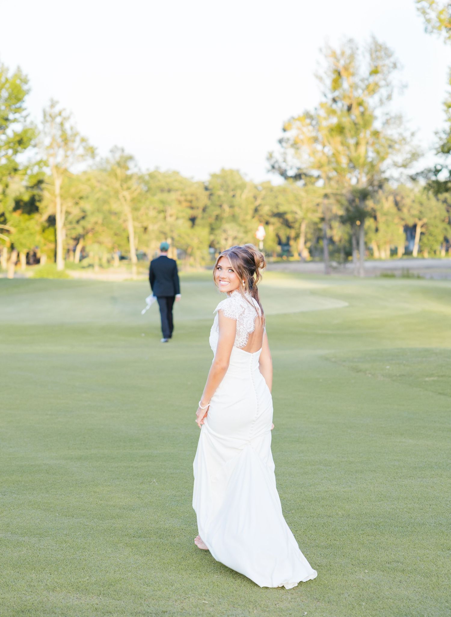 Bride in white open-back gown smiles over shoulder on golf course as groom walks away in background.