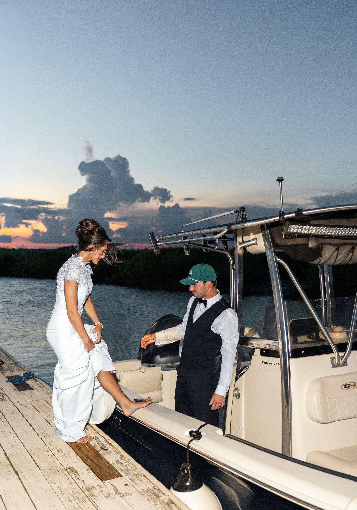 Bride in white gown stepping onto a boat at sunset, assisted by a man in formal attire at a waterfront dock.
