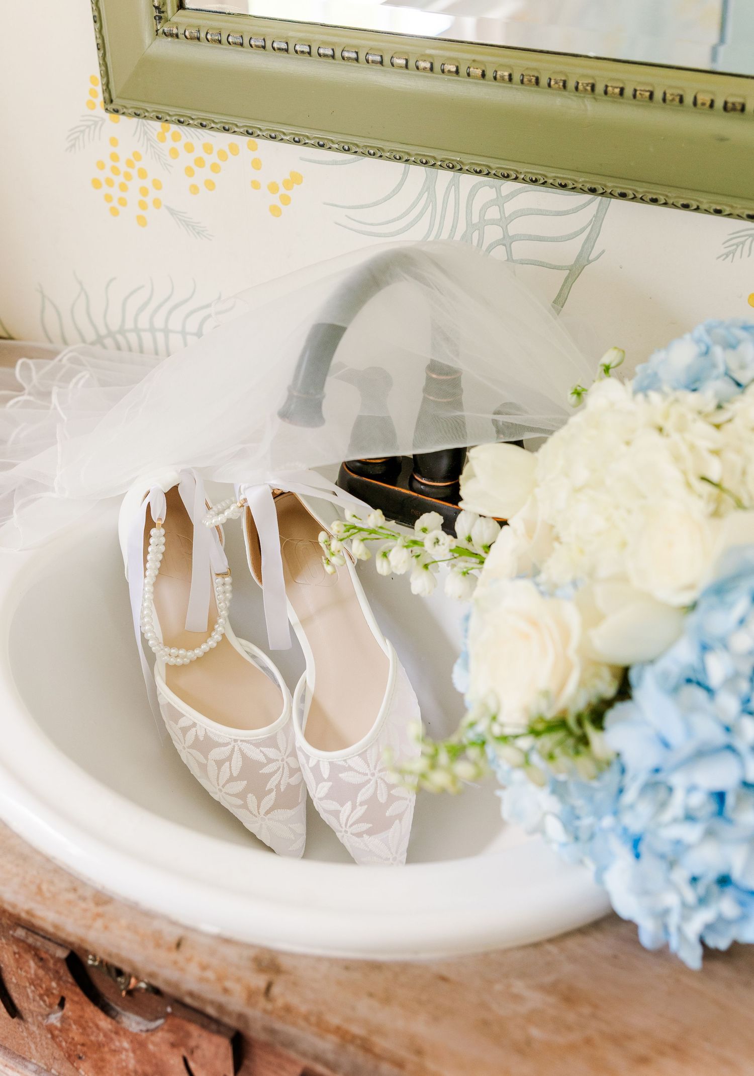 White lace bridal heels with pearl ankle strap displayed in sink beside blue hydrangea bouquet.