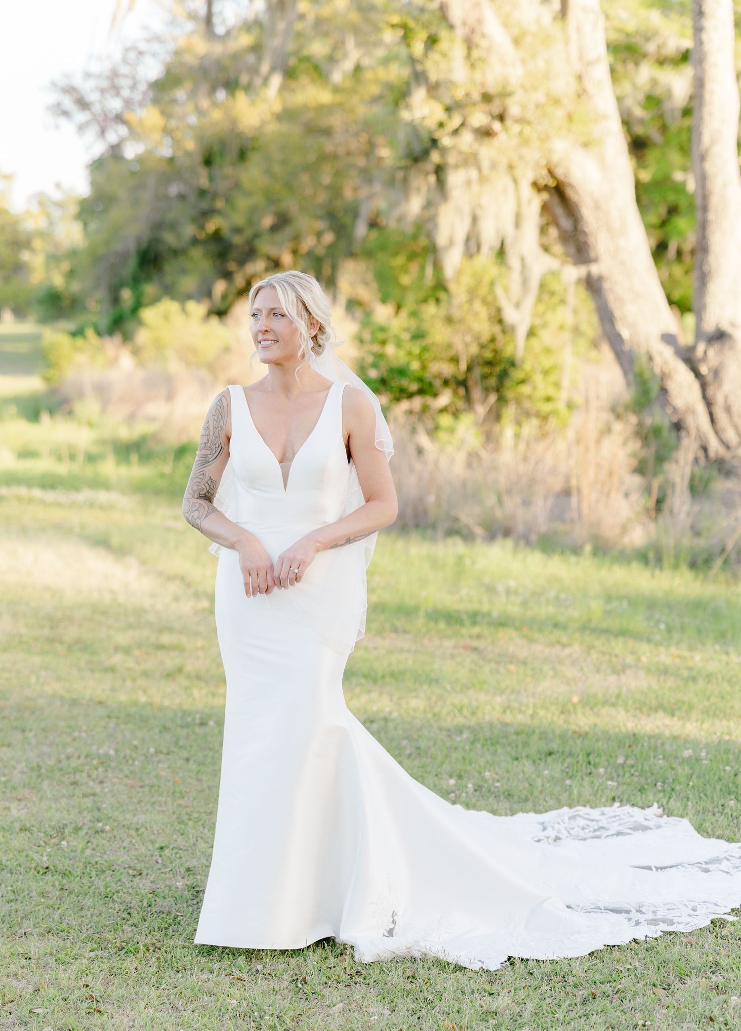 Bride in fitted white mermaid wedding dress with train smiling in sunlit outdoor setting.