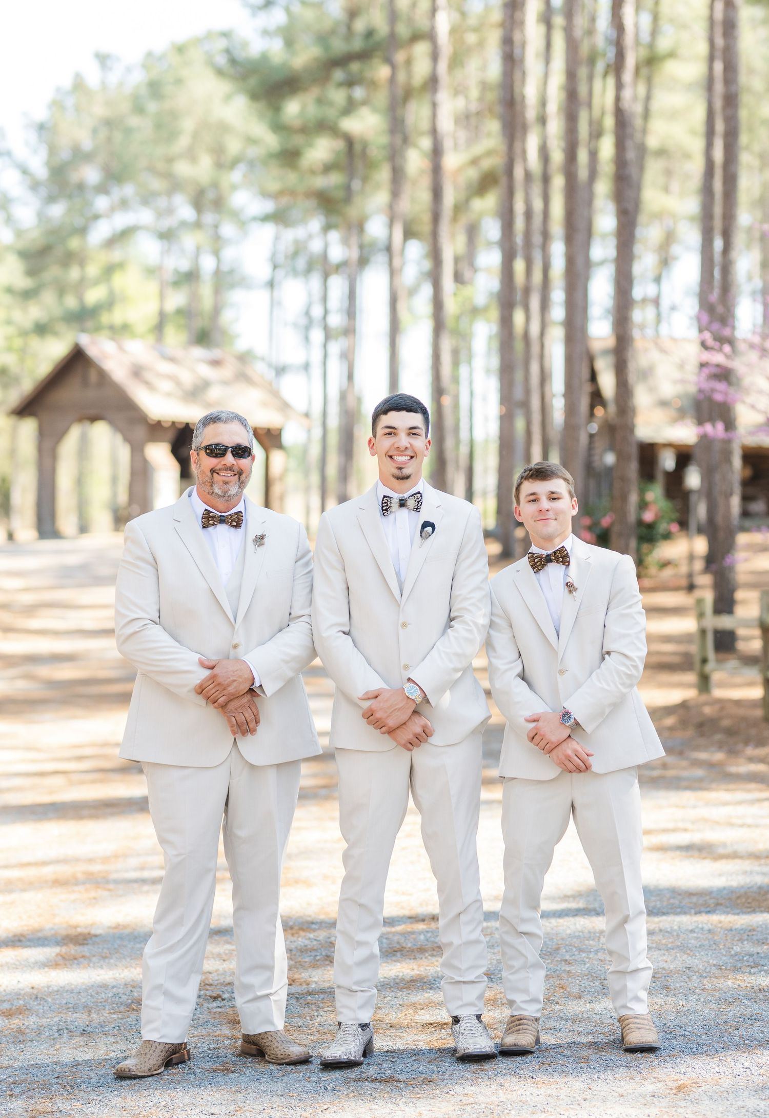 Groom and two groomsmen in matching cream suits with bow ties posing in pine tree setting.