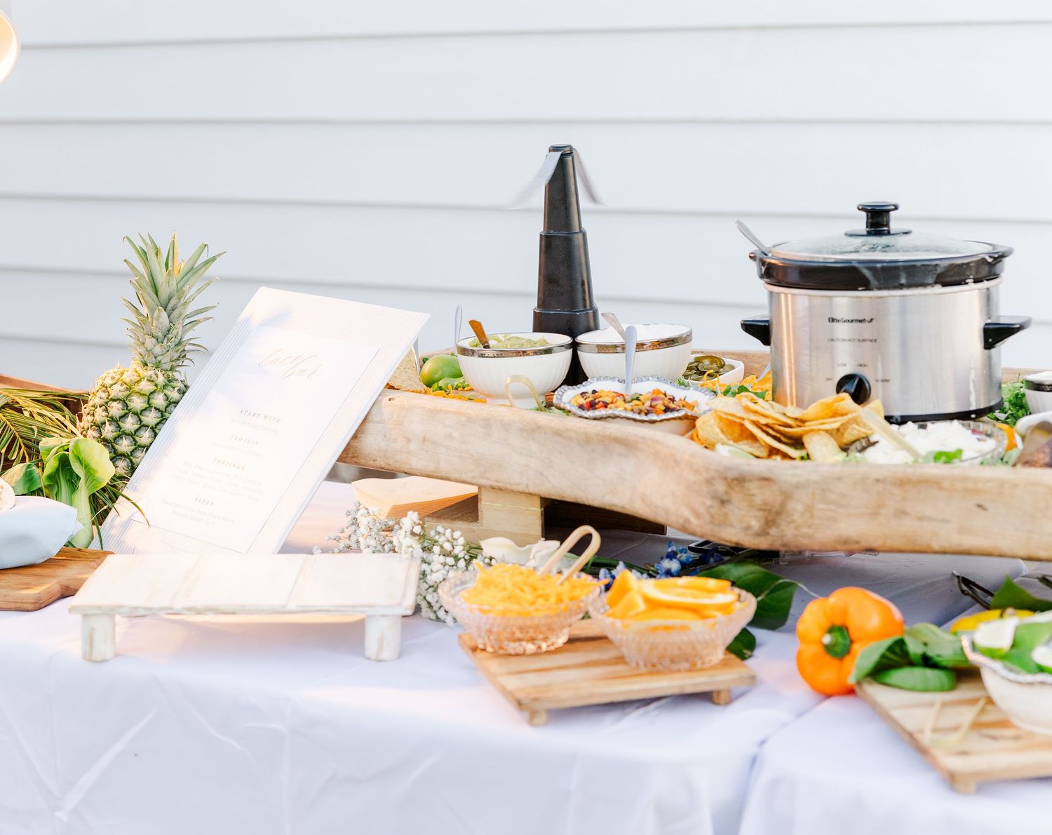 Rustic wooden grazing table with pineapple, chips, dips, and a slow cooker at an outdoor wedding reception.